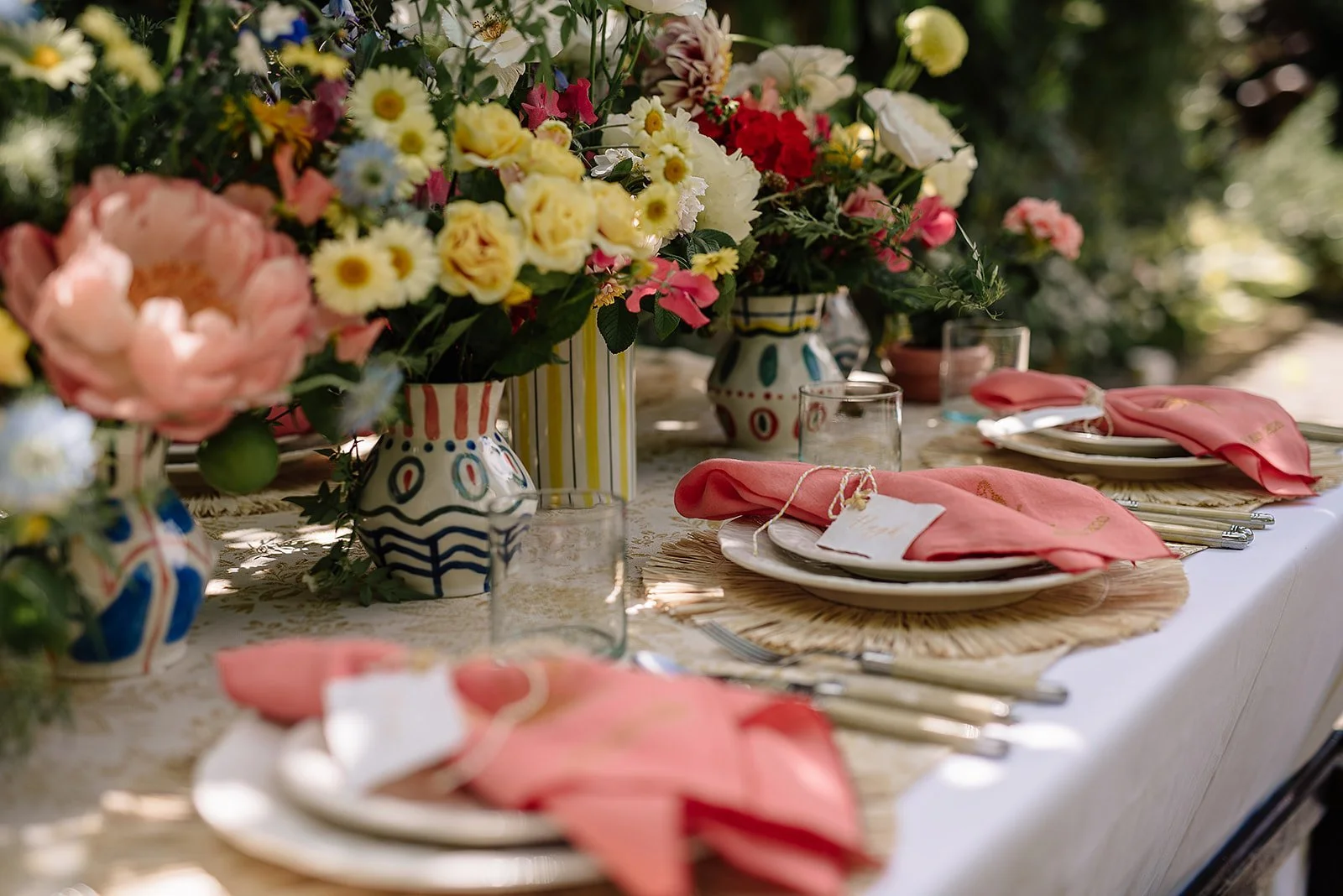A table set for an outdoor meal with colorful floral centerpieces in decorative vases, pink napkins, clear glassware, and white plates on woven placemats under dappled sunlight.