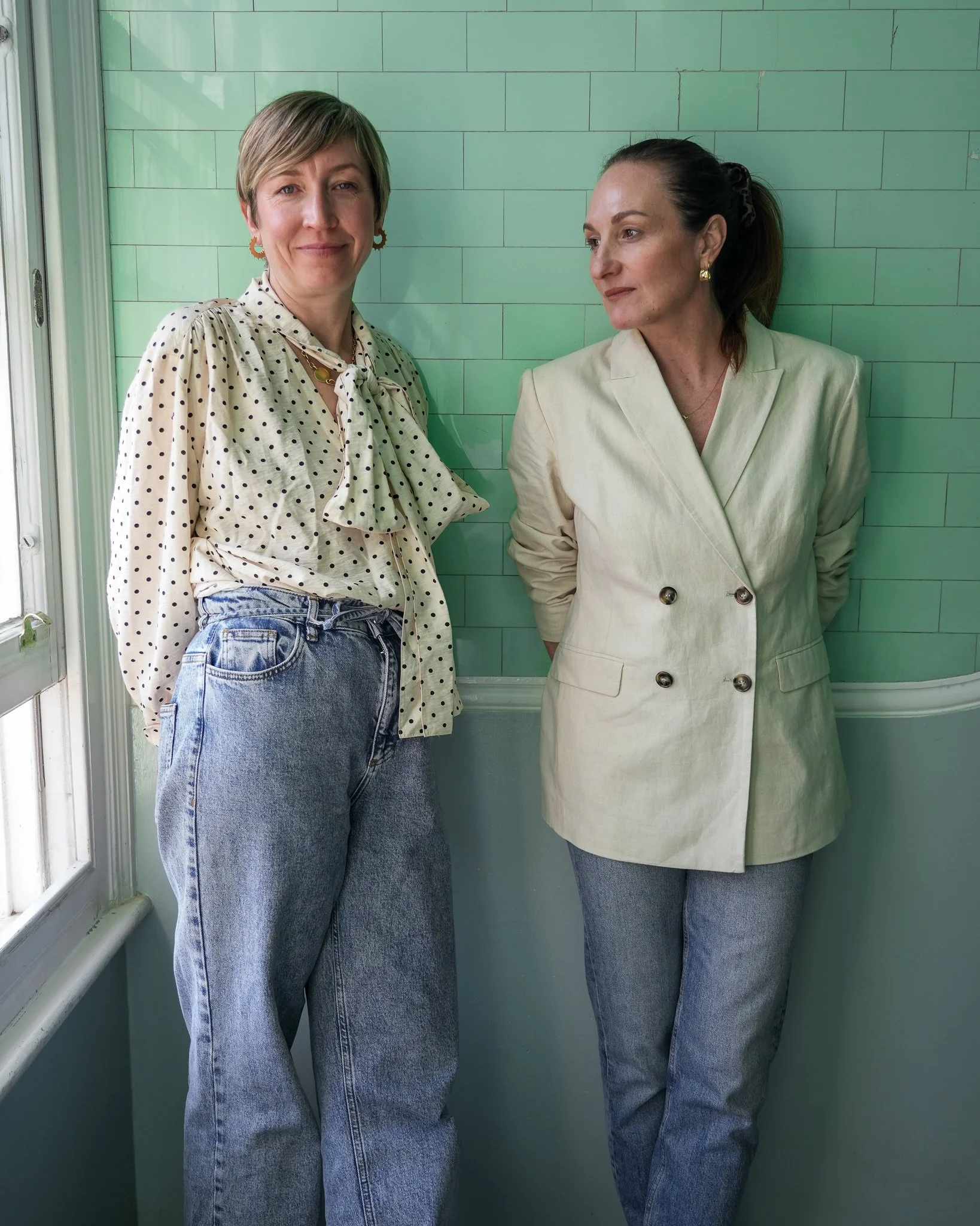 Two women with short and long hair standing against a green tiled wall, wearing cream-colored jackets and jeans, looking at each other.