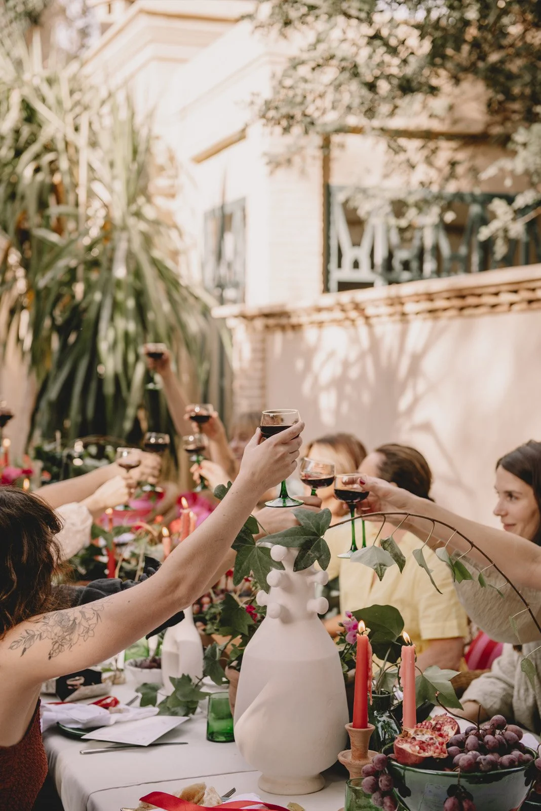 Group of women gathered outdoors, raising glasses of red wine for a toast at a table decorated with candles, flowers, and greenery.
