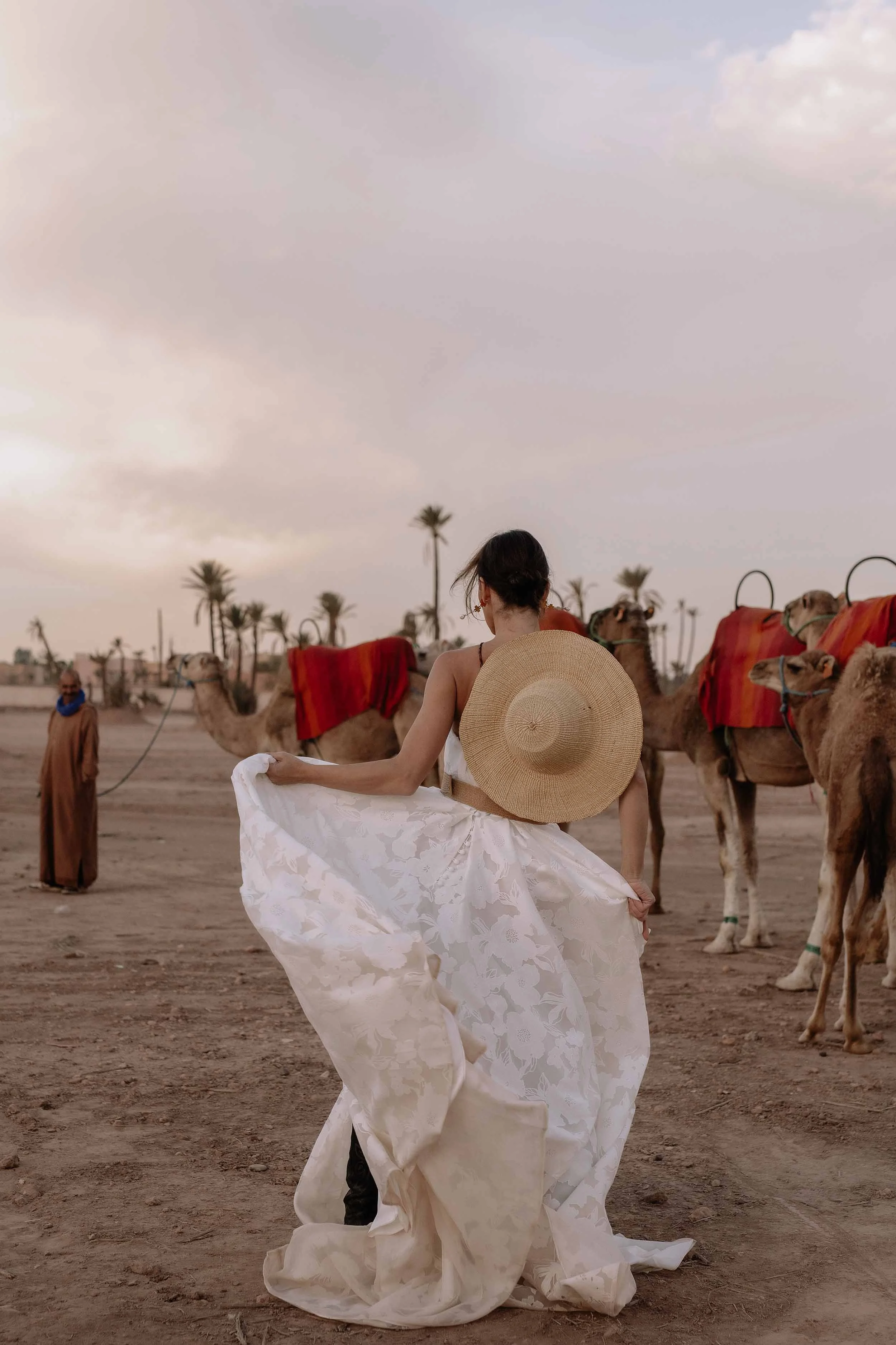 A woman in a white dress holding a wide-brimmed hat, standing in front of camels against a desert backdrop with palm trees and a cloudy sky.