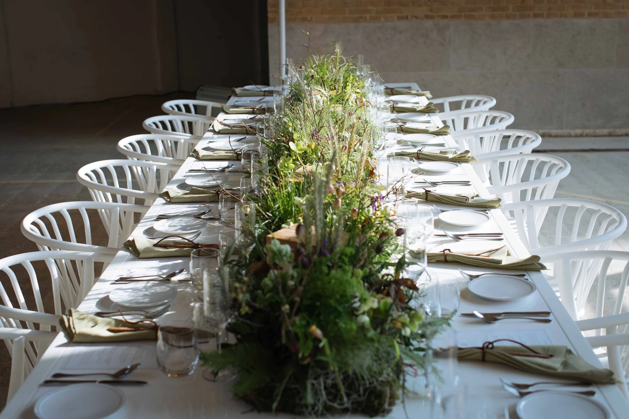 A long dining table set for a formal event, with white chairs, neatly folded napkins, glassware, and a lush green floral centerpiece.
