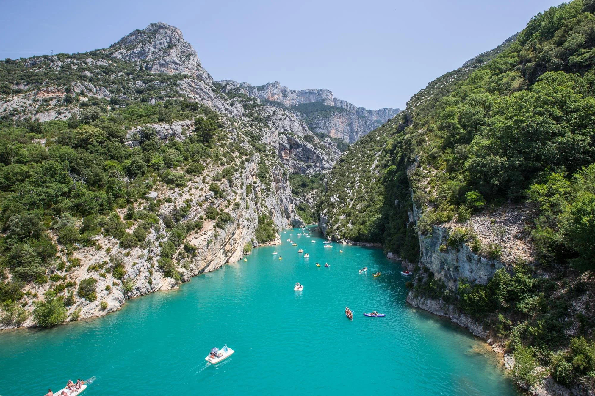 Vue d'une rivière turquoise entourée de falaises rocheuses et de forêts verdoyantes, avec plusieurs bateaux flottant sur l'eau, sous un ciel clair.