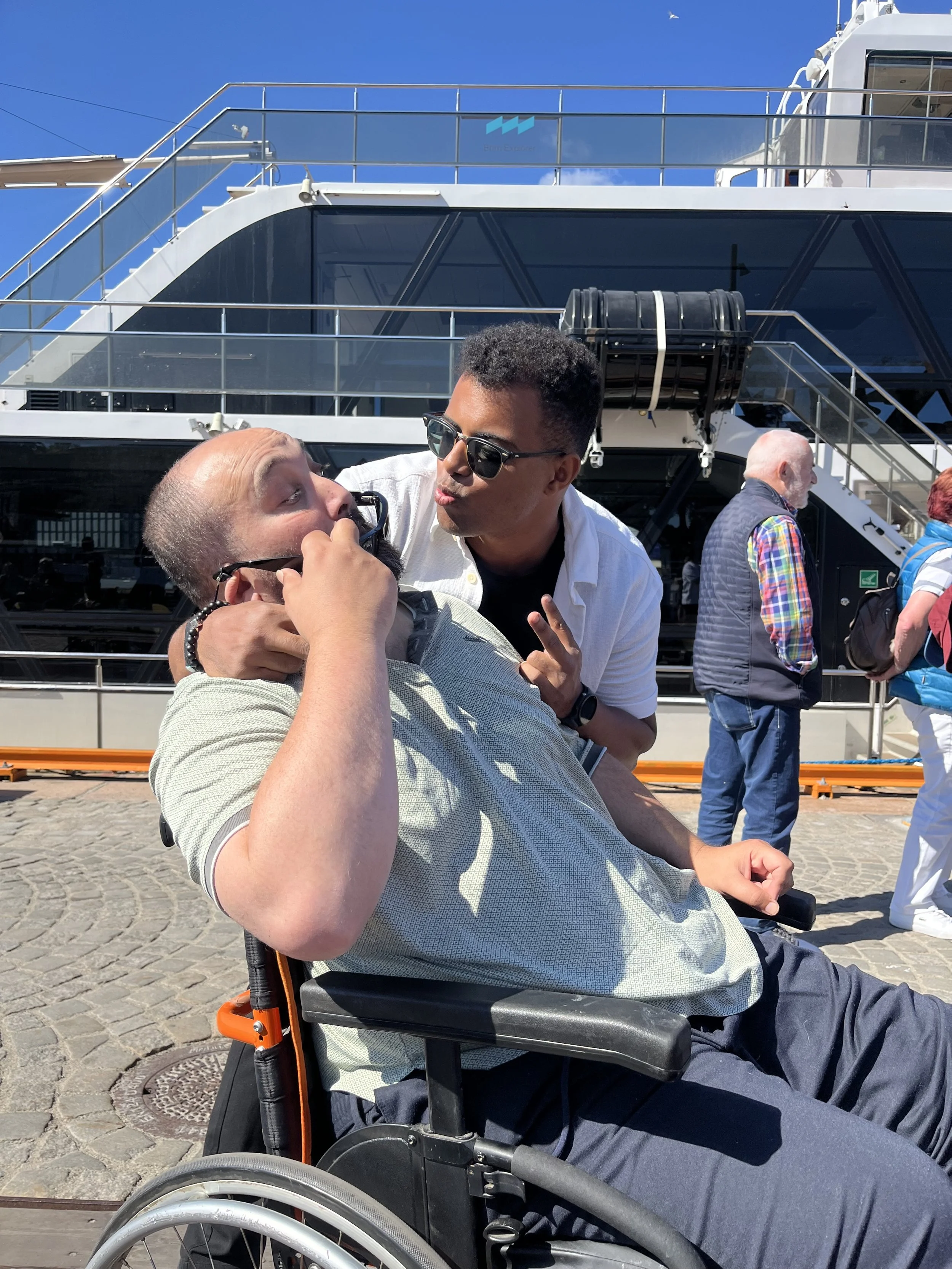Une personne en fauteuil roulant en conversation avec une autre personne devant un grand navire de croisière, en plein jour.