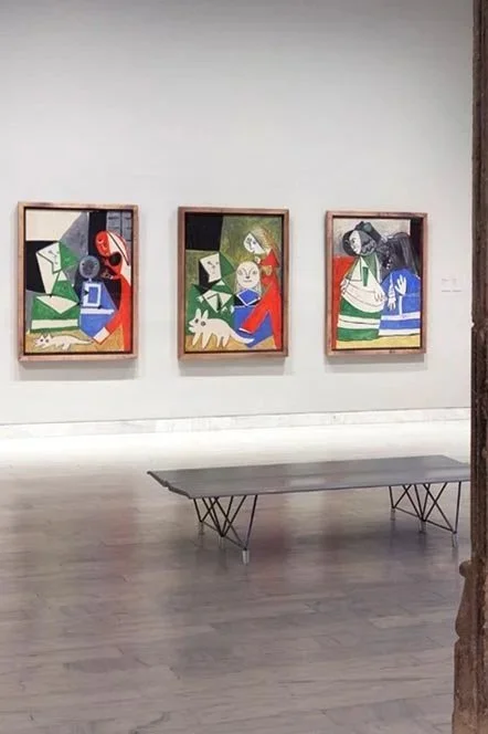 Just Travel met en avant trois peintures modernes illustrant des personnages abstraits, accrochées sur un mur blanc dans une galerie d'art, avec un banc au premier plan.