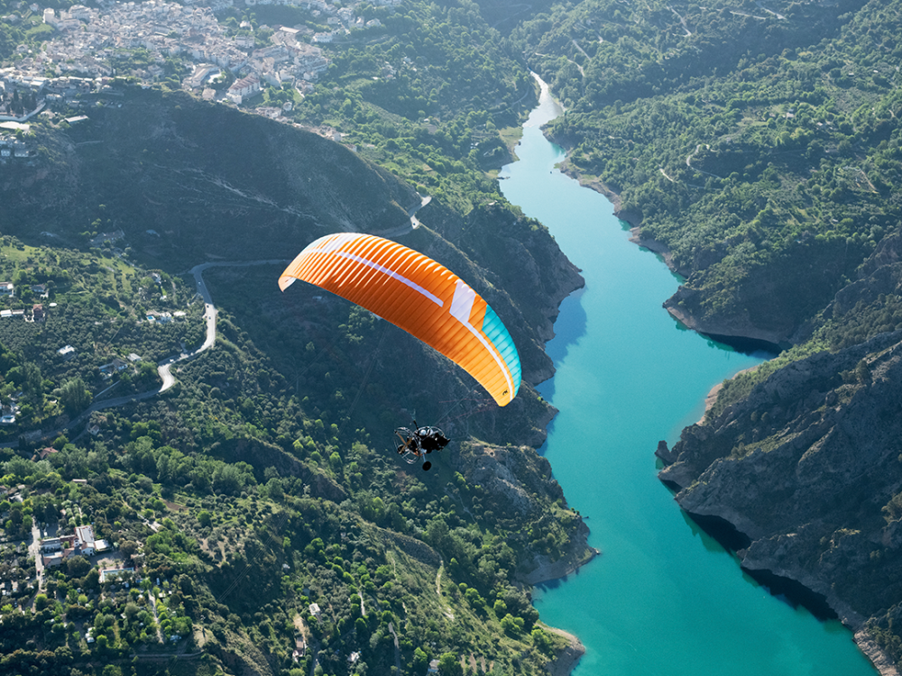 Parapente orange survolant une vallée verdoyante avec un fleuve turquoise, entourée de montagnes. Une activité accessible aux personnes en situation de handicap.