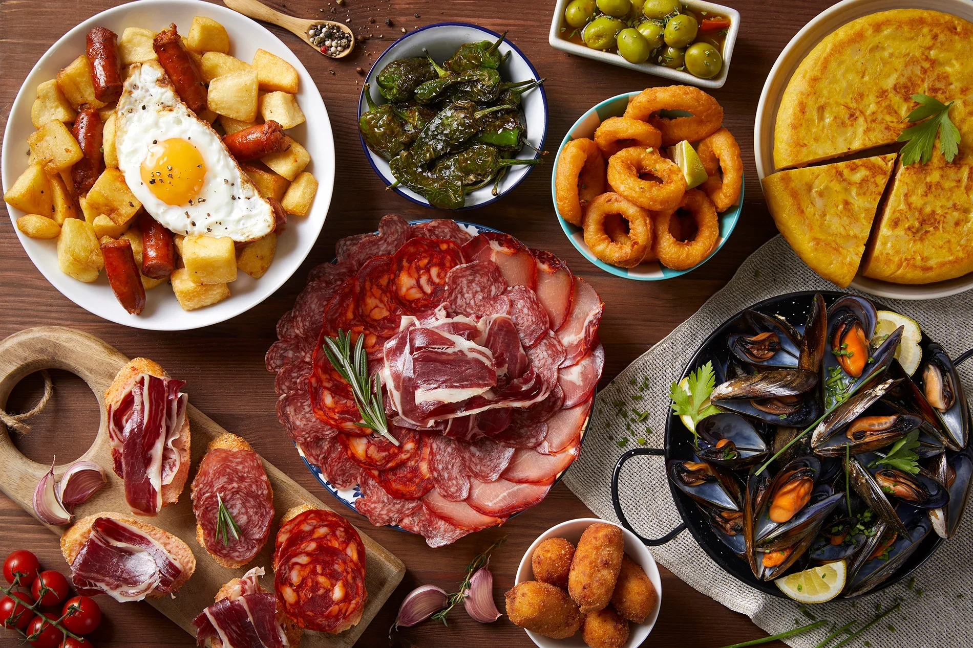 Just Travel dévoile un assortiment de plats méditerranéens comprenant du jambon, charcuterie, moules, frites avec œuf au plat, salade de piments, fondue ou tortilla, calamars frits, roquette, et olives vertes, tous présentés sur une table en bois.