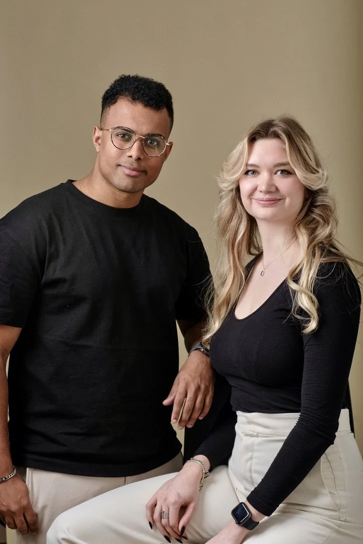 Deux personnes, un homme et une femme, posent ensemble contre un fond beige. L'homme porte un t-shirt noir, des lunettes, et a la peau foncée, la femme porte un haut noir, des cheveux blonds, et une montre intelligente au poignet gauche.