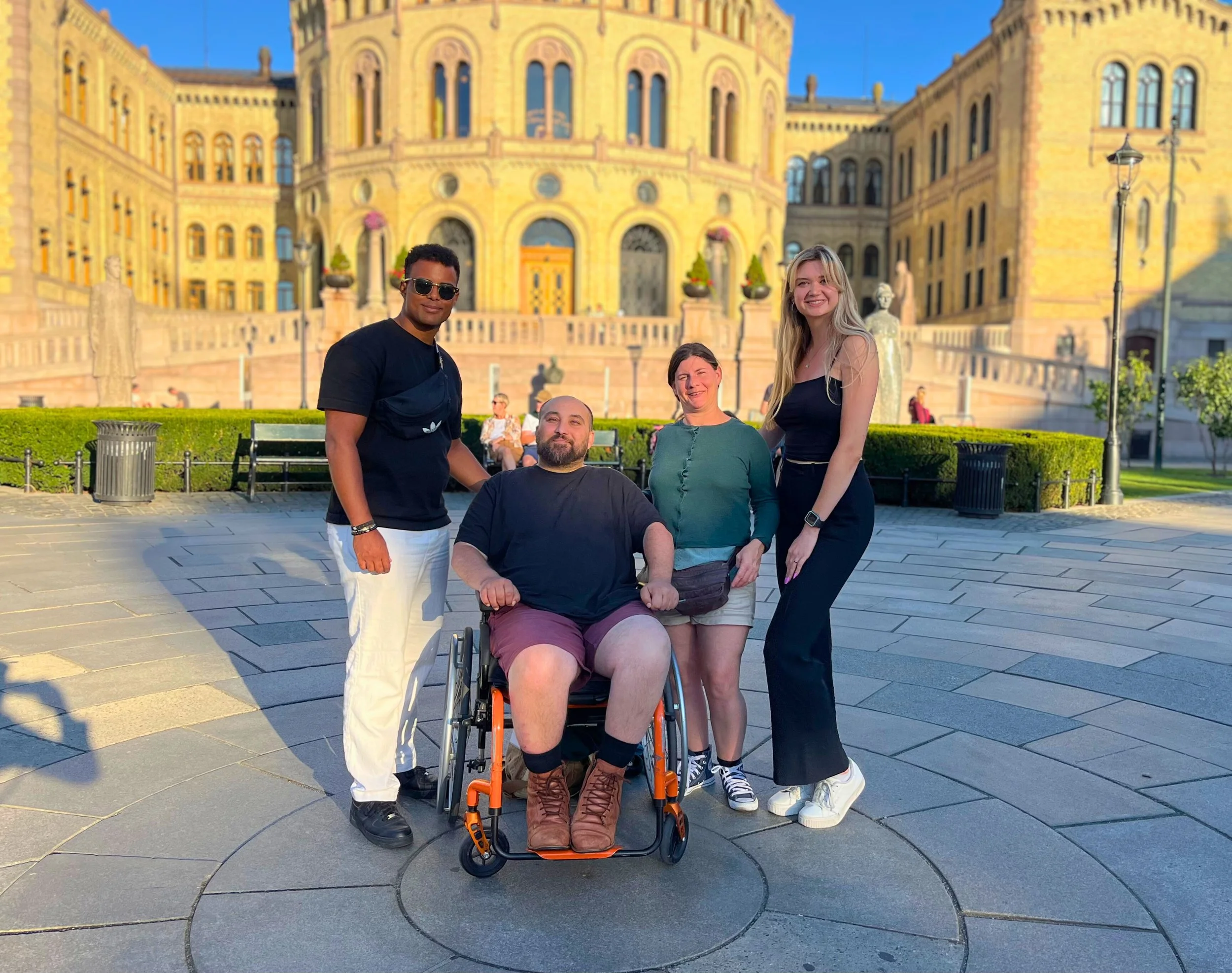 Quatre personnes posent devant un bâtiment ancien et imposant. Parmi elles, un homme en fauteuil roulant, deux femmes et un homme portant des vêtements décontractés. La scène se déroule en plein jour, dans un espace ouvert pavé avec des bancs et des lampadaires, ensoleillé et agréable.