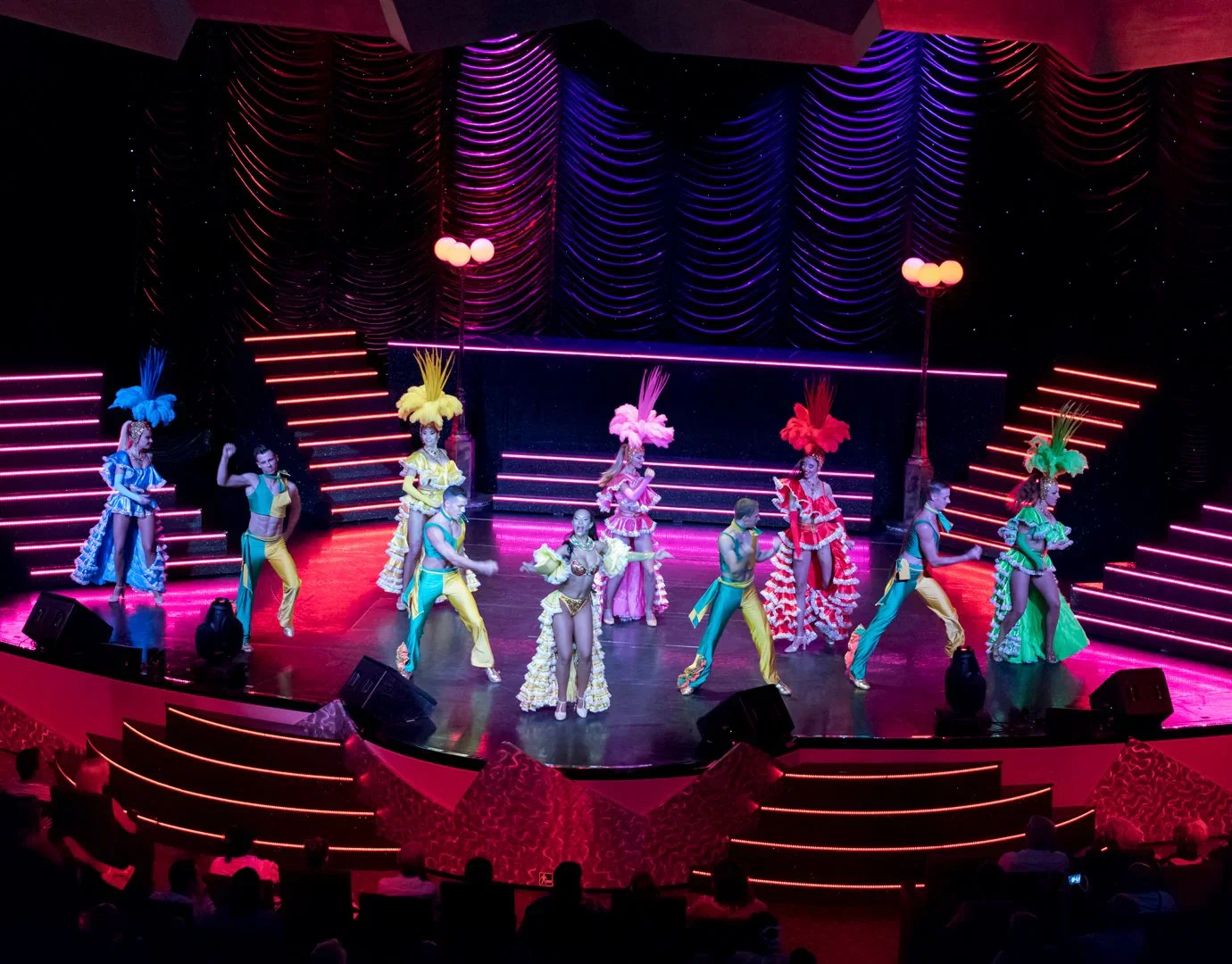 Just Travel vous montre un spectacle de danse avec des artistes en costumes colorés sur scène éclairée de lumières roses et violettes, dans une salle avec un public en arrière-plan.