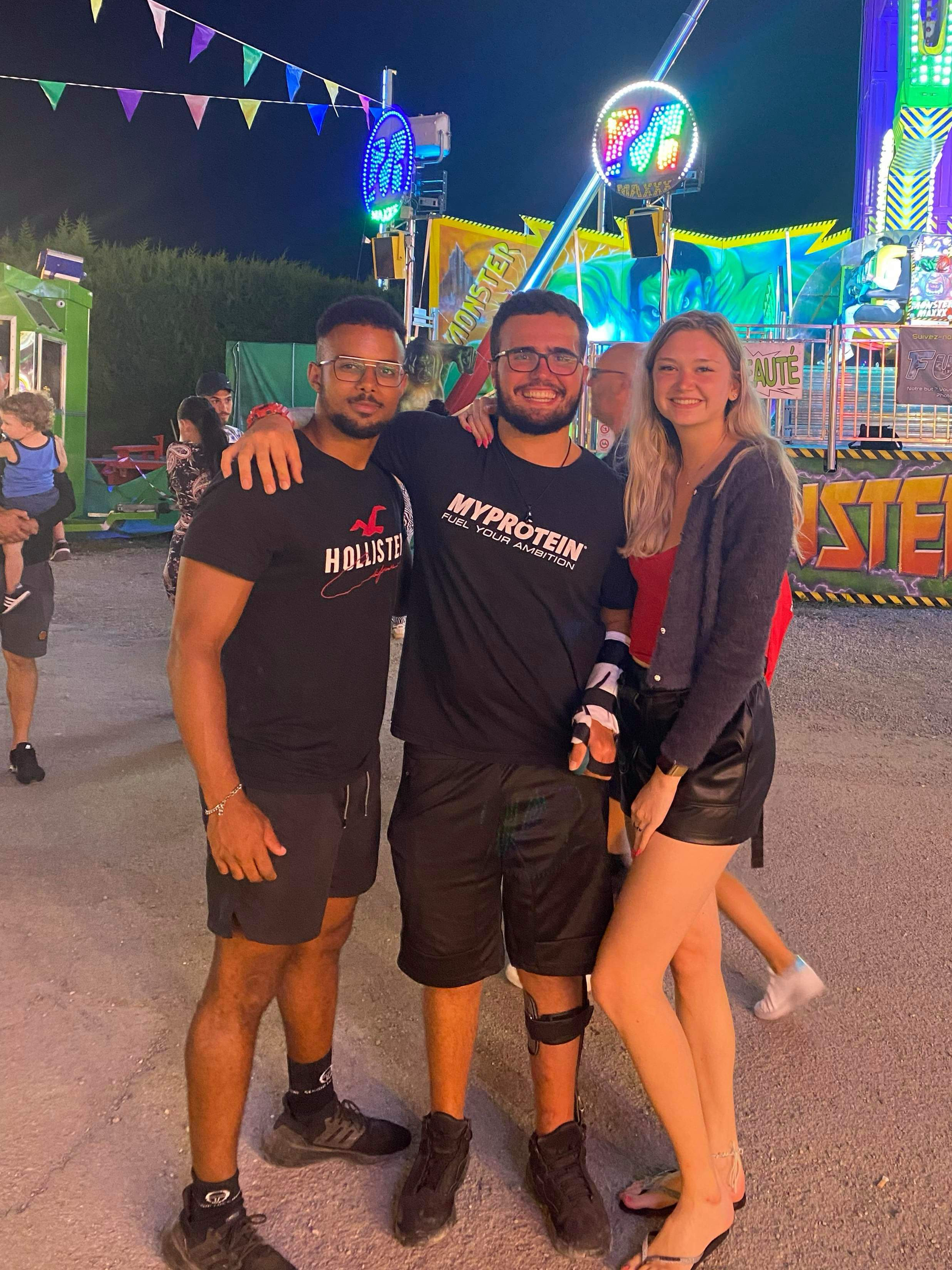 Trois jeunes adultes souriants posent ensemble devant une fête foraine la nuit, avec des attractions illuminées en arrière-plan.
