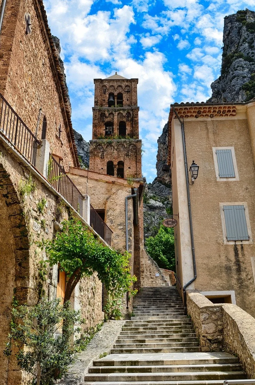 Vue d'une ruelle pavée avec des escaliers menant à une vieille église en pierre avec un clocher, entourée de bâtiments en pierres et de montagnes rocheuses, sous un ciel bleu avec des nuages blancs.