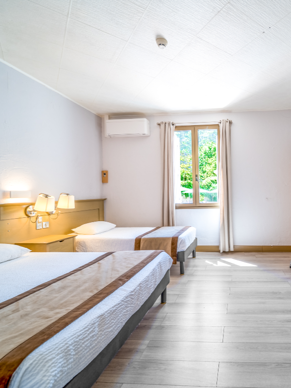 Chambre d'hôtel avec deux lits, une fenêtre laissant entrer la lumière naturelle, un climatiseur, et une décoration simple.