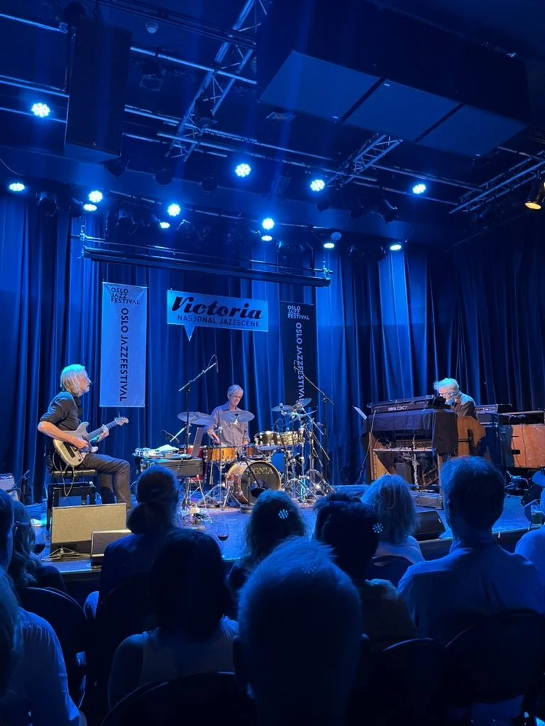 Un groupe de musiciens jouant sur scène lors d'un festival de jazz à Oslo, avec un public assis en face. La scène est éclairée en bleu, avec des panneaux indiquant "Victoria" et "Nazional Jazz Scene".