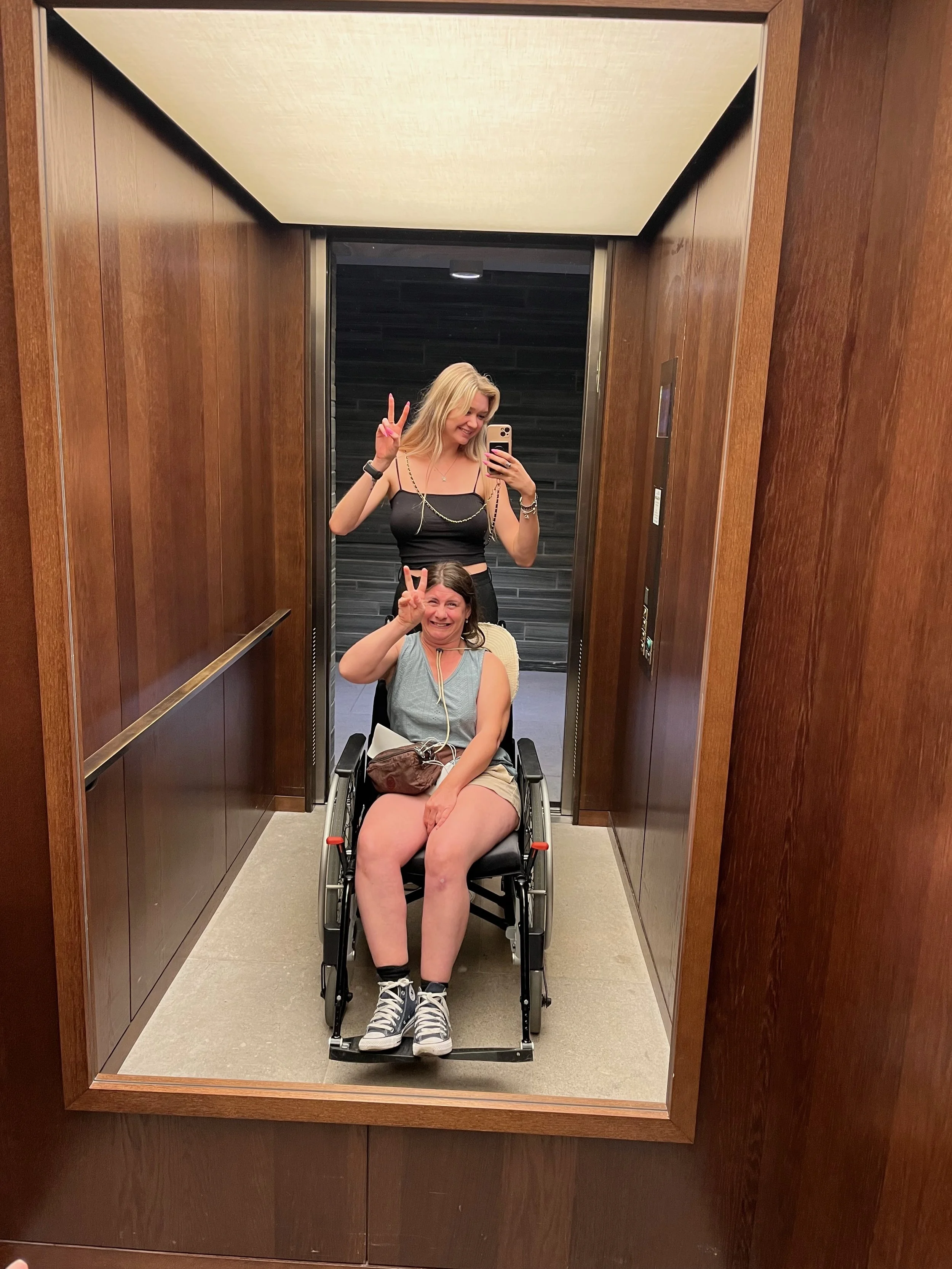 Deux jeunes femmes prennent un selfie dans un ascenseur avec un miroir. La femme assise est en fauteuil roulant, portant un t-shirt gris et des shorts. La femme derrière, debout, porte un haut noir avec des détails en or et fait un signe de paix.