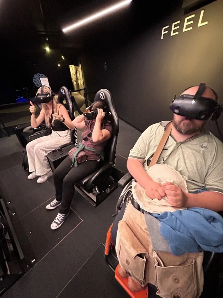 Quatre personnes assises dans une pièce sombre, portant des casques de réalité virtuelle. Trois sont des femmes et un est un homme. Les femmes donnent un pouce en l'air, l'homme tient un objet rond.