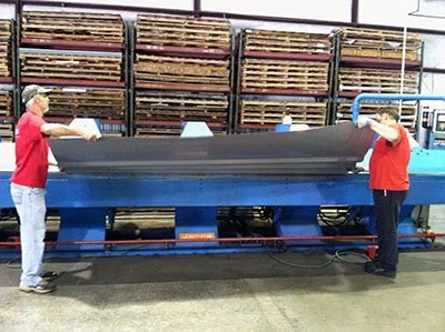 Two workers in red shirts and jeans operating a large sheet fabric or paper cutting machine in a warehouse.