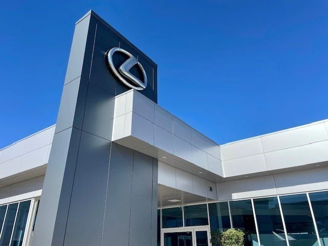 Exterior of a Lexus car dealership with a large Lexus logo on a tall sign against a blue sky.