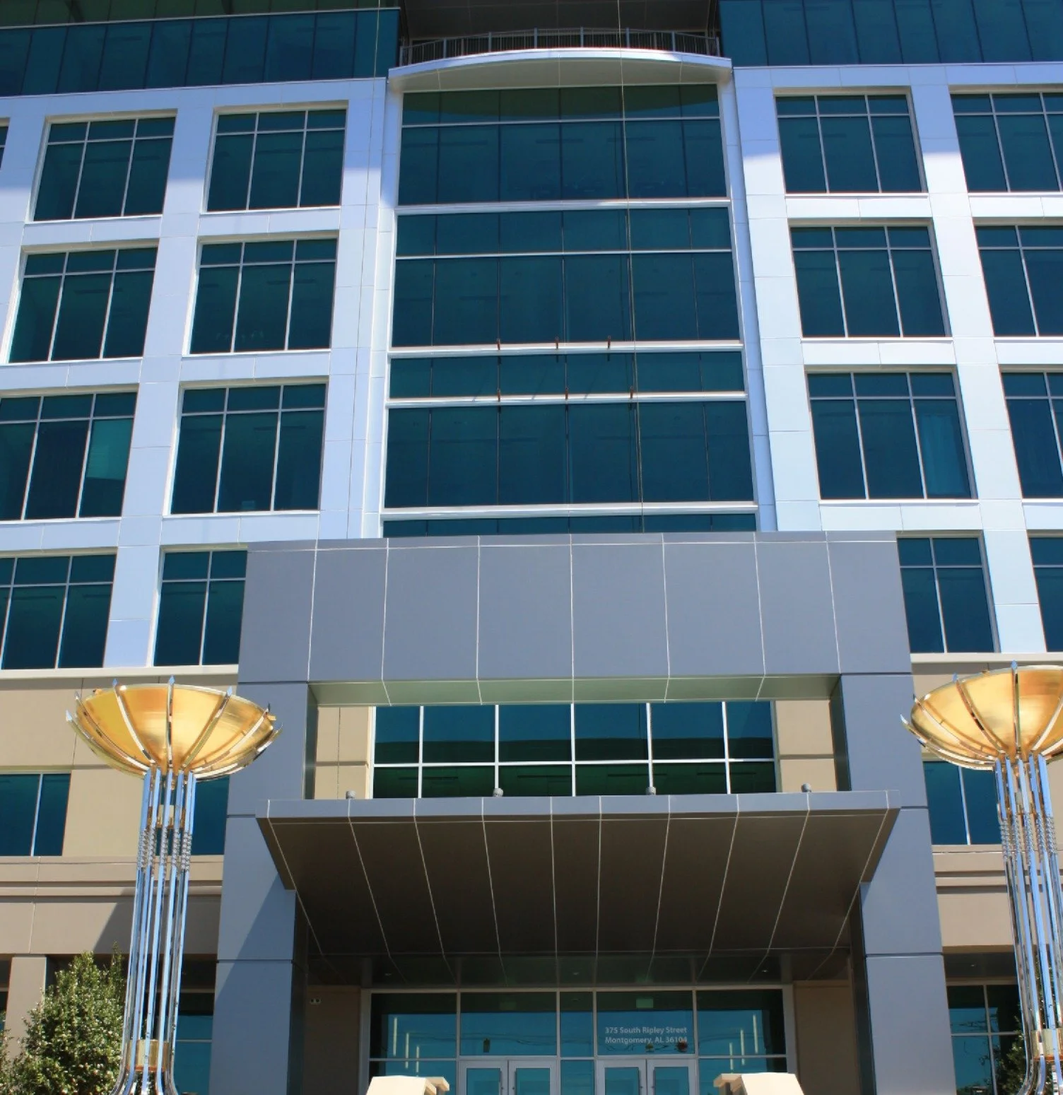 Front view of a modern multi-story office building with large glass windows and a gray and beige facade, featuring two decorative gold-colored street lamps at the entrance.