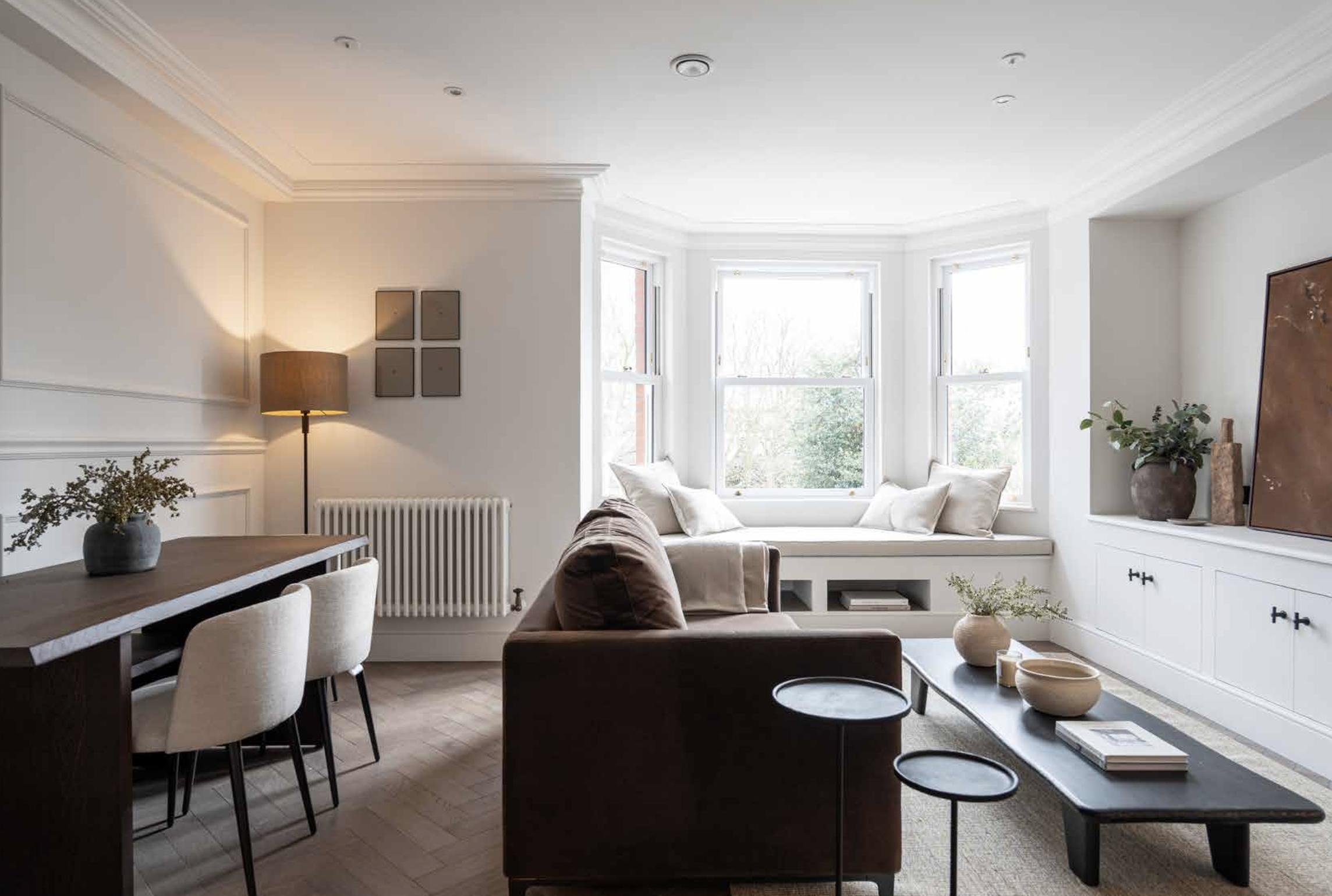 Bright living room with white walls, bay window seat with pillows, brown sofa, black coffee table with decorative bowls, beige rug, and white cabinets with decor, including plants and artwork.