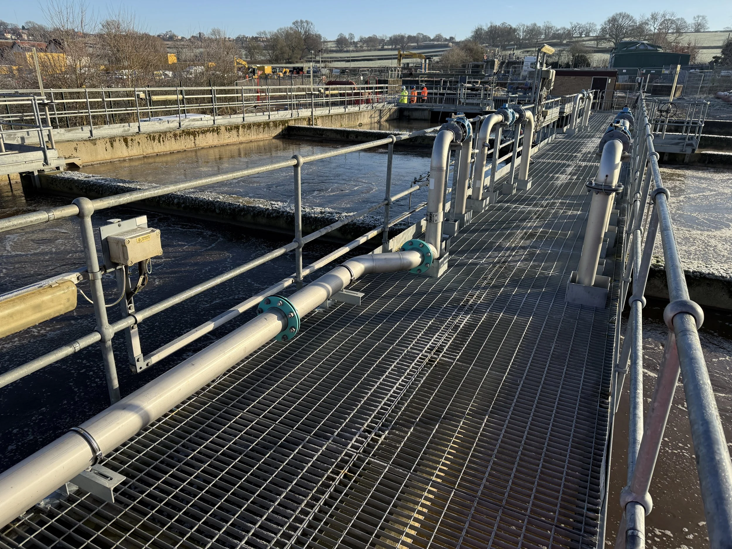 Photograph of an industrial water treatment facility showing metal walkways, pipes, and large tanks with water, with construction workers in the background.
