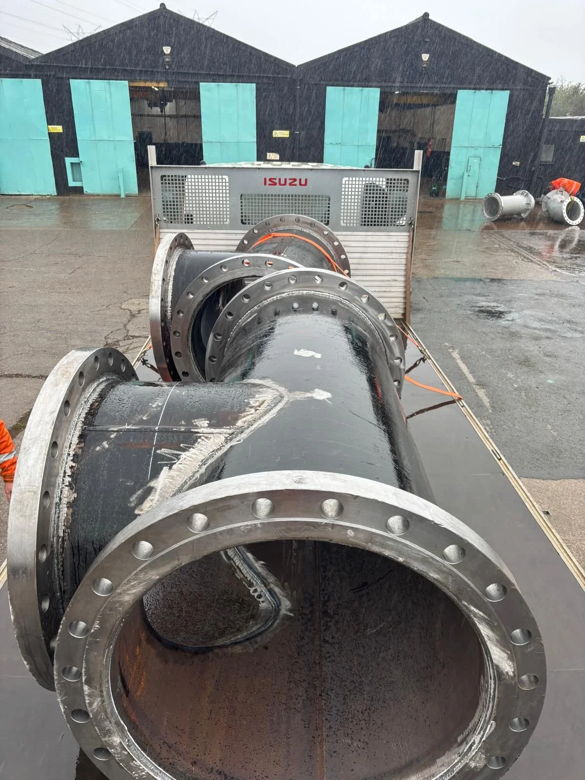 Large black industrial pipe with flanged ends on a flat surface, rain pouring down, industrial building in background.