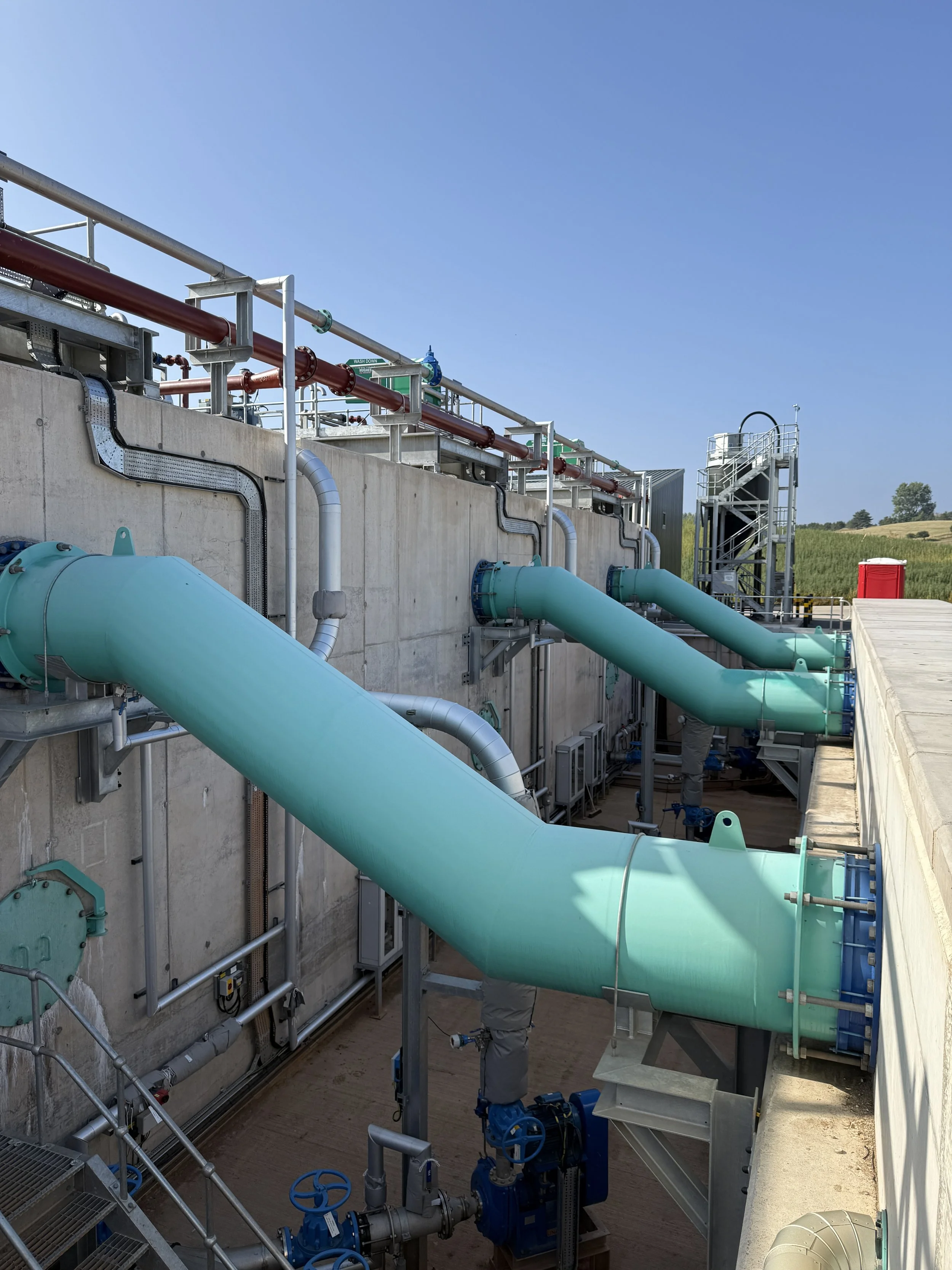 View of industrial pipes, valves, and machinery outside on a sunny day, with a staircase and green field in the background.