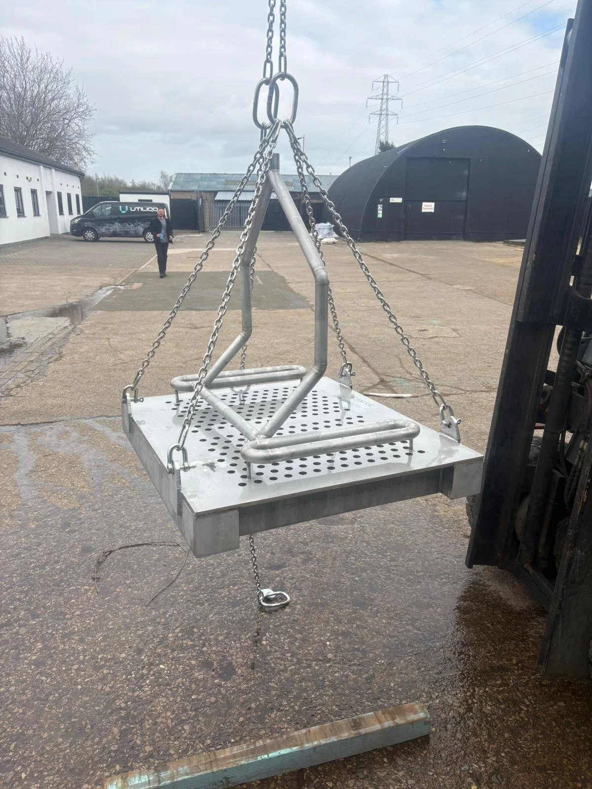 A metal swing seat being lifted by a forklift vehicle outdoors with a person walking in the background.