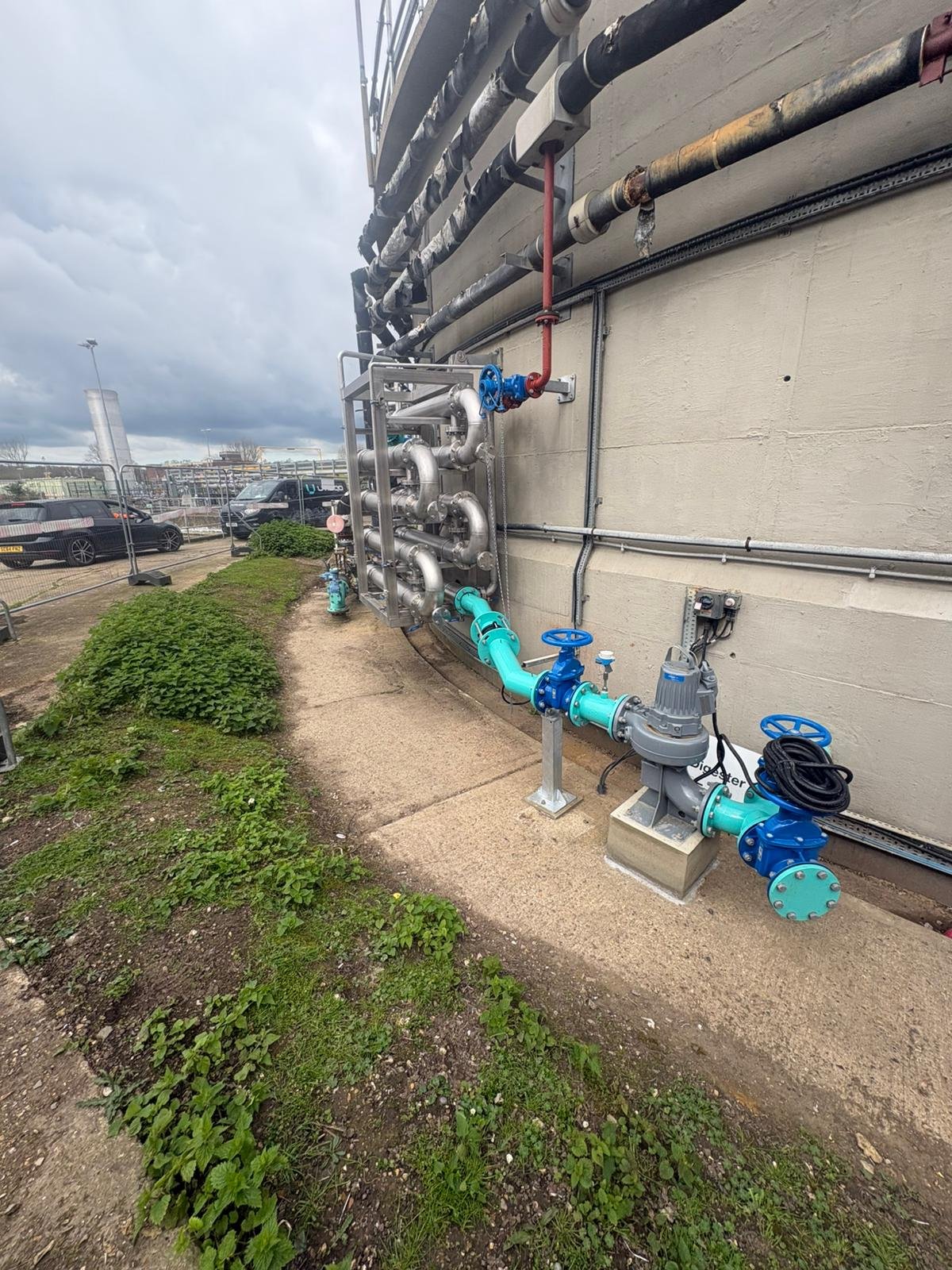 Outdoor industrial piping system with blue, gray, and metallic pipes and valves attached to a beige building wall. Parking lot and cloudy sky in the background.