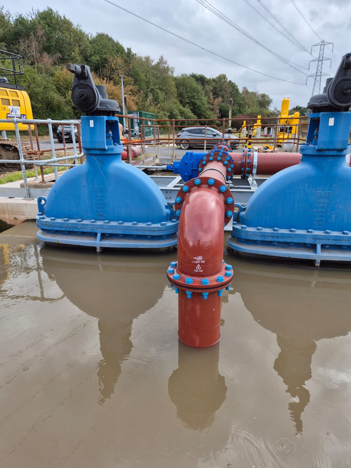 Large industrial valves and pipes at a water treatment or utility site, with a water body in the foreground and green hills and sky in the background.