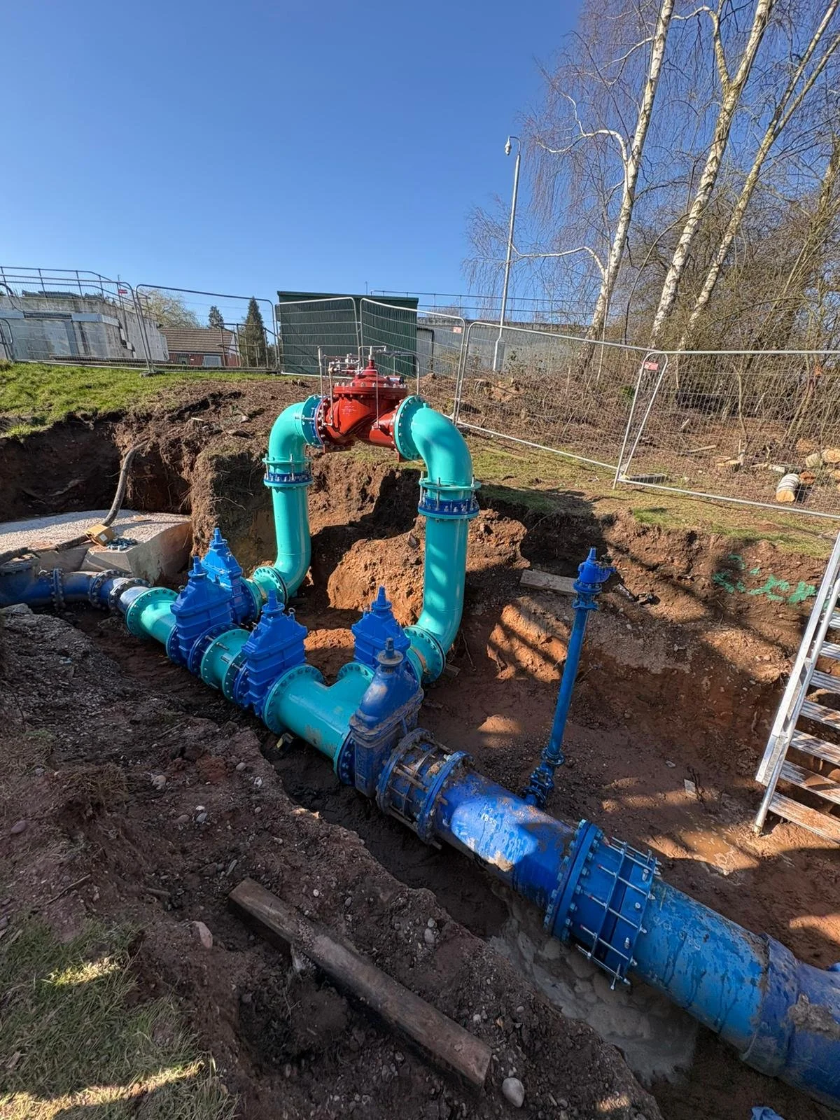 Installation of large water pipes underground, depicting blue and red pipes, valves, and fittings, with an open trench and a fence in the background under a clear blue sky.