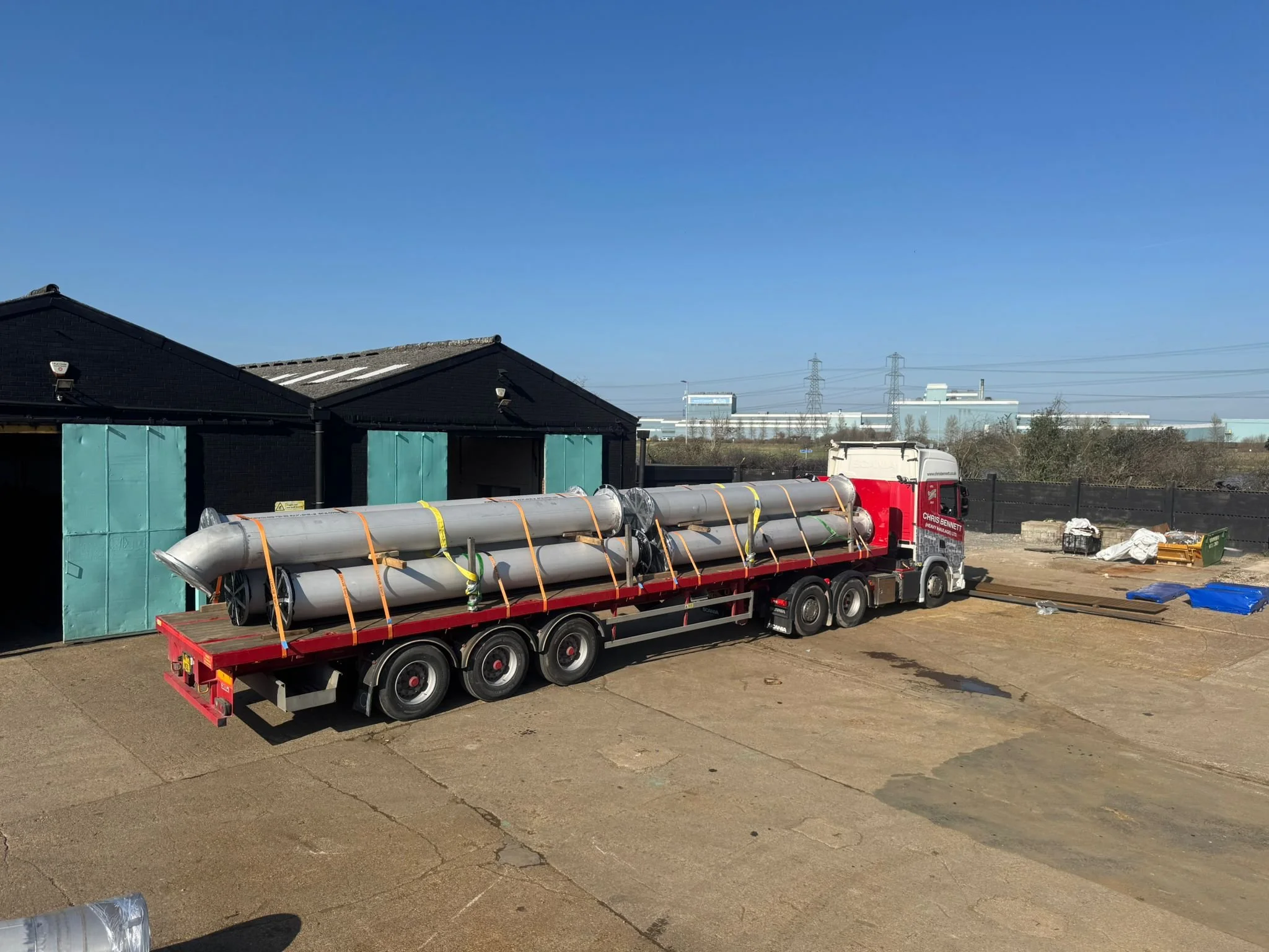 A flatbed truck carrying large pipes, parked in an industrial or construction area with a building, power lines, and clear blue sky in the background.