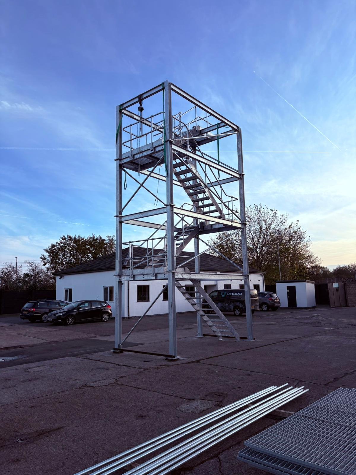 Metal construction framework with stairs, set up outdoors on a paved area, with cars, small white buildings, and trees in the background.
