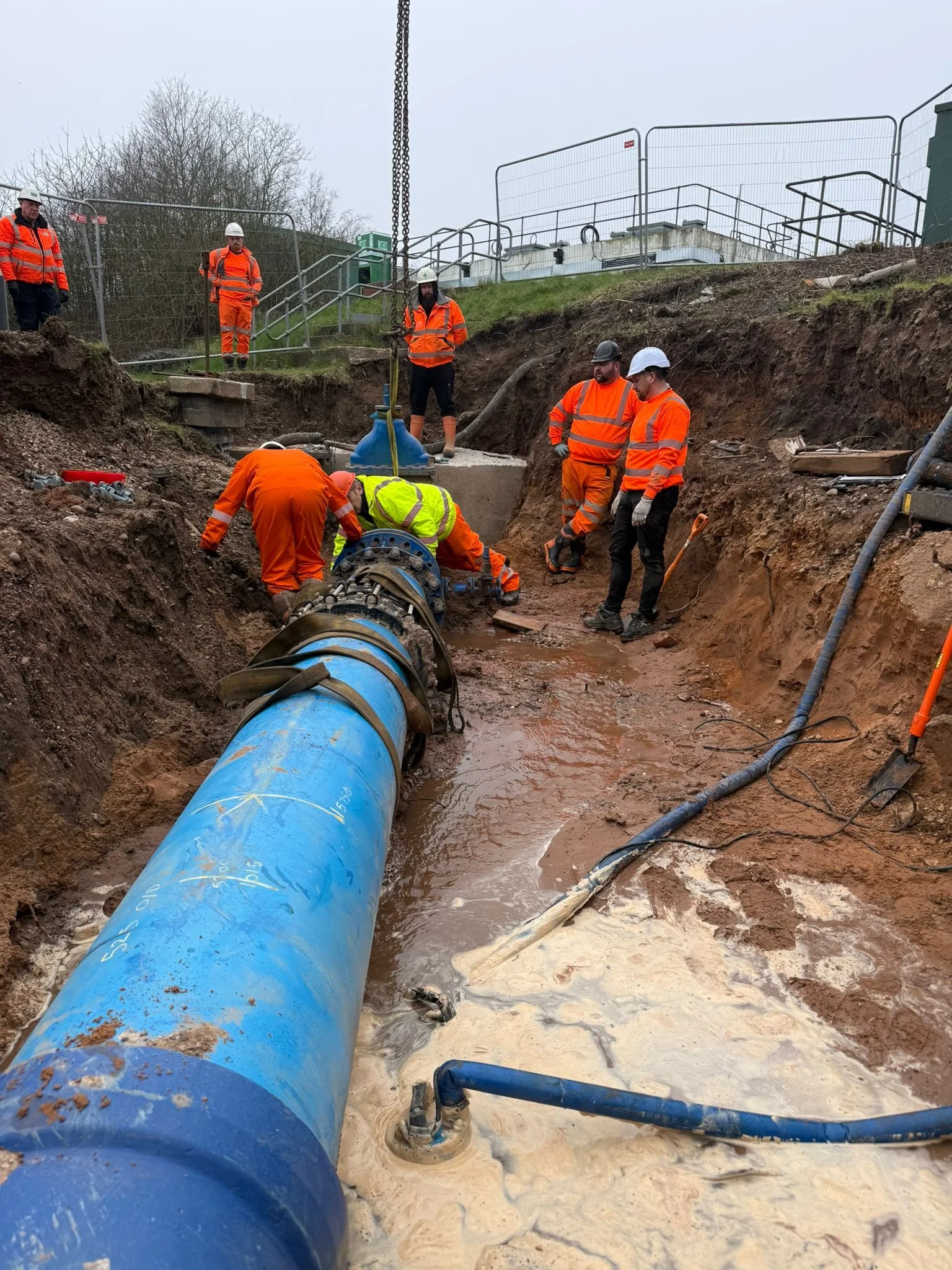 Workers in orange and yellow safety gear repairing a large blue pipeline at a construction site, with muddy ground and industrial equipment.
