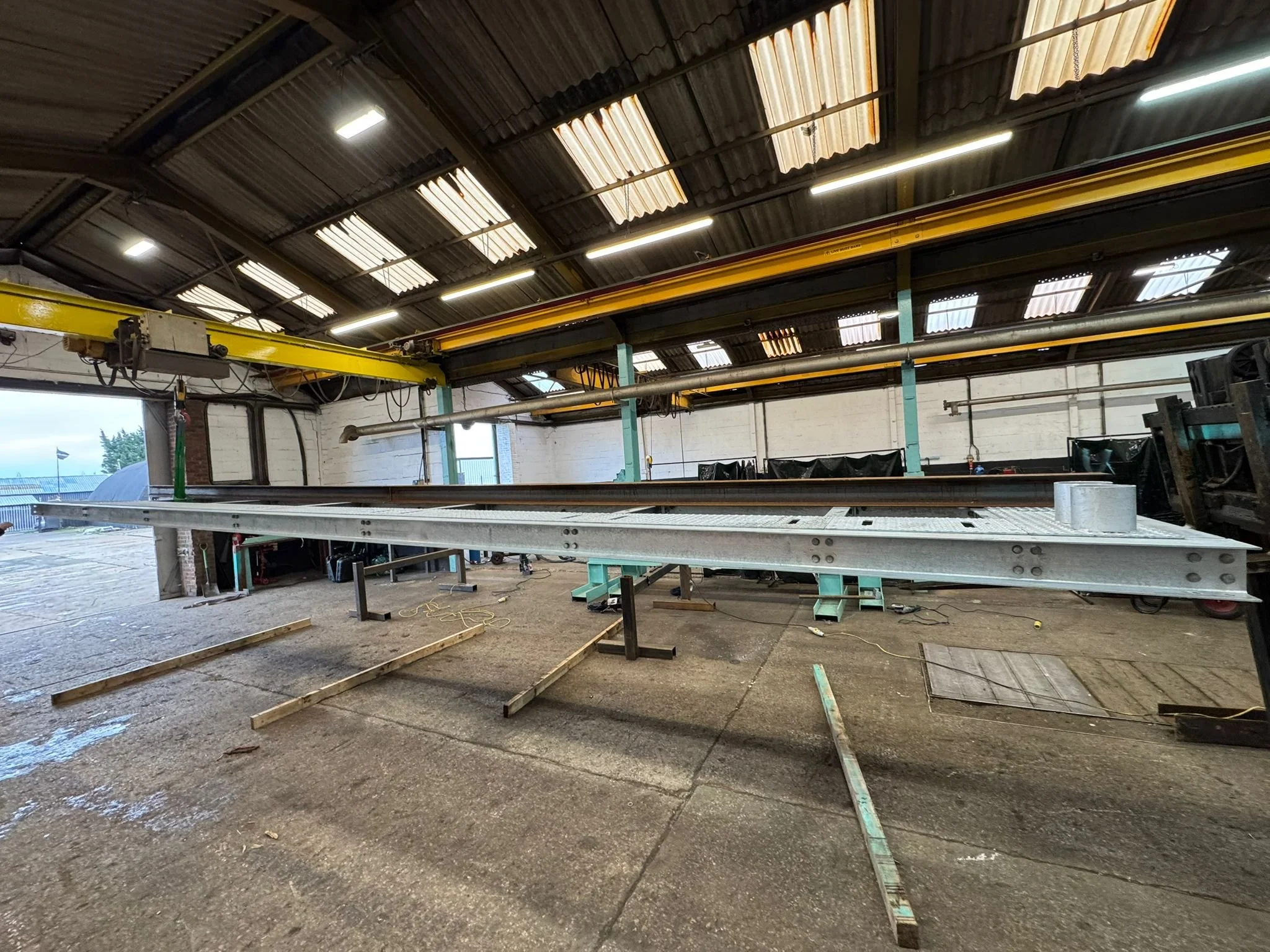 Inside a workshop or garage with a metal roof and fluorescent lighting, showing metal beams and a large yellow overhead crane, with workbenches and equipment below.