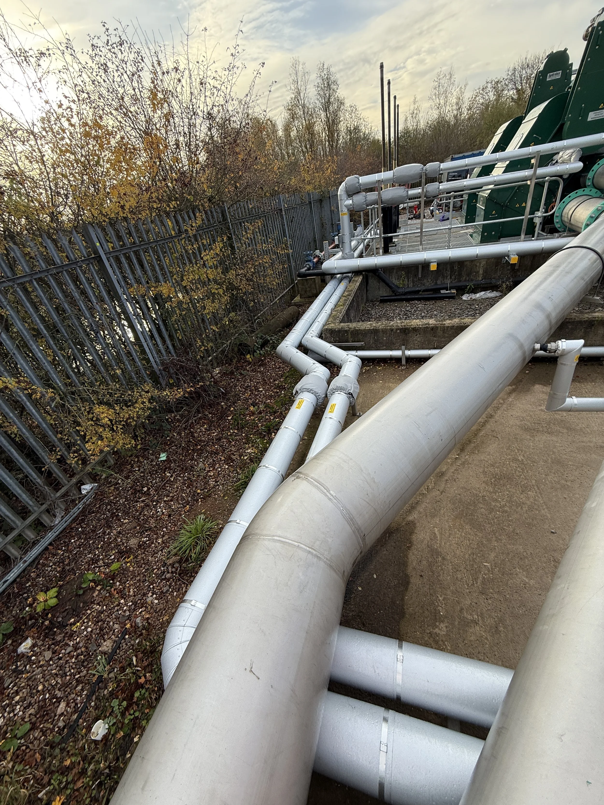 Large industrial pipes and equipment outdoors next to a protective metal fence and trees with autumn leaves, under a cloudy sky.