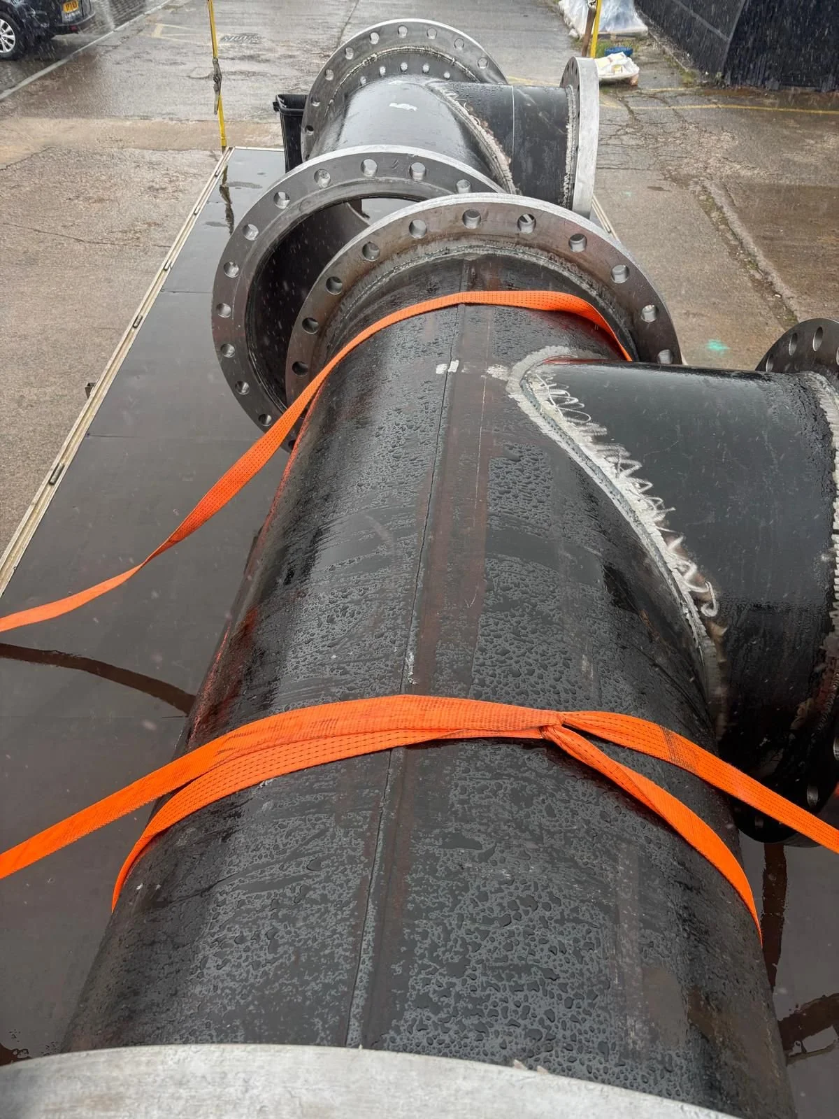 View from the back of a large black pipe with flanged ends, secured with orange straps, on a flatbed truck in a rainy outdoor setting.