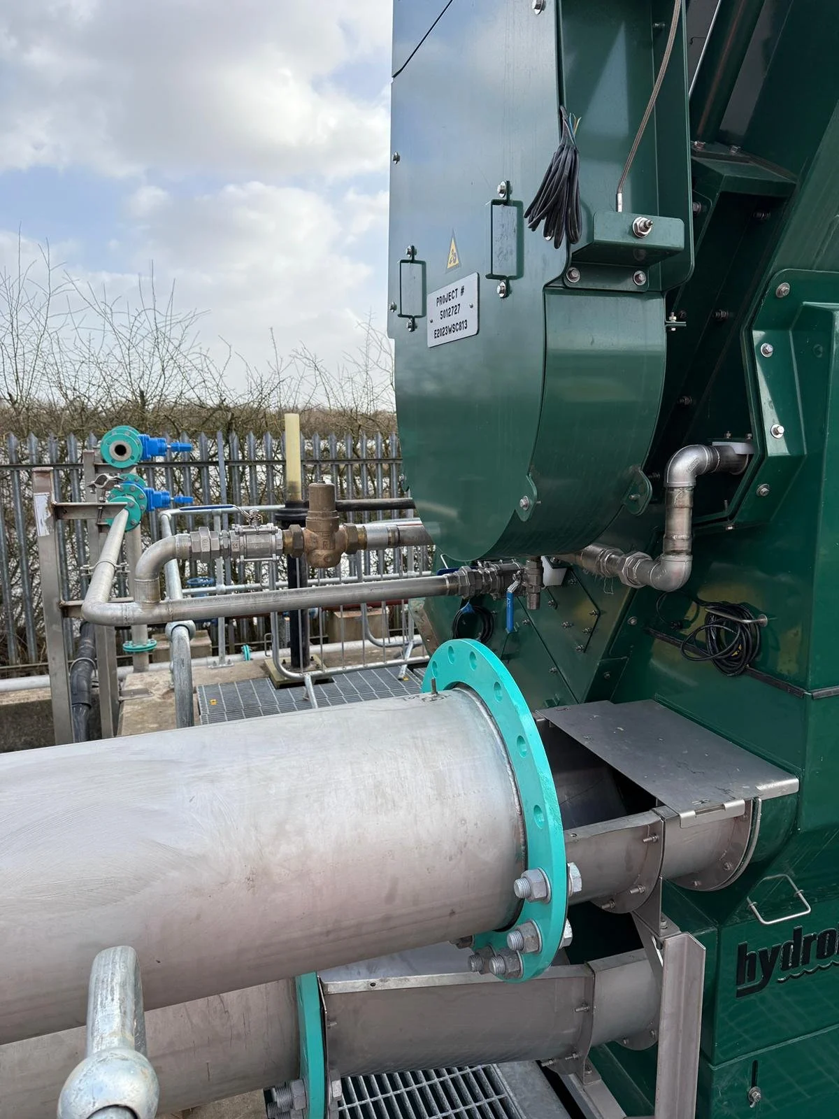 Industrial equipment with pipes, valves, and a large green metal box, outdoors with cloudy sky in the background.