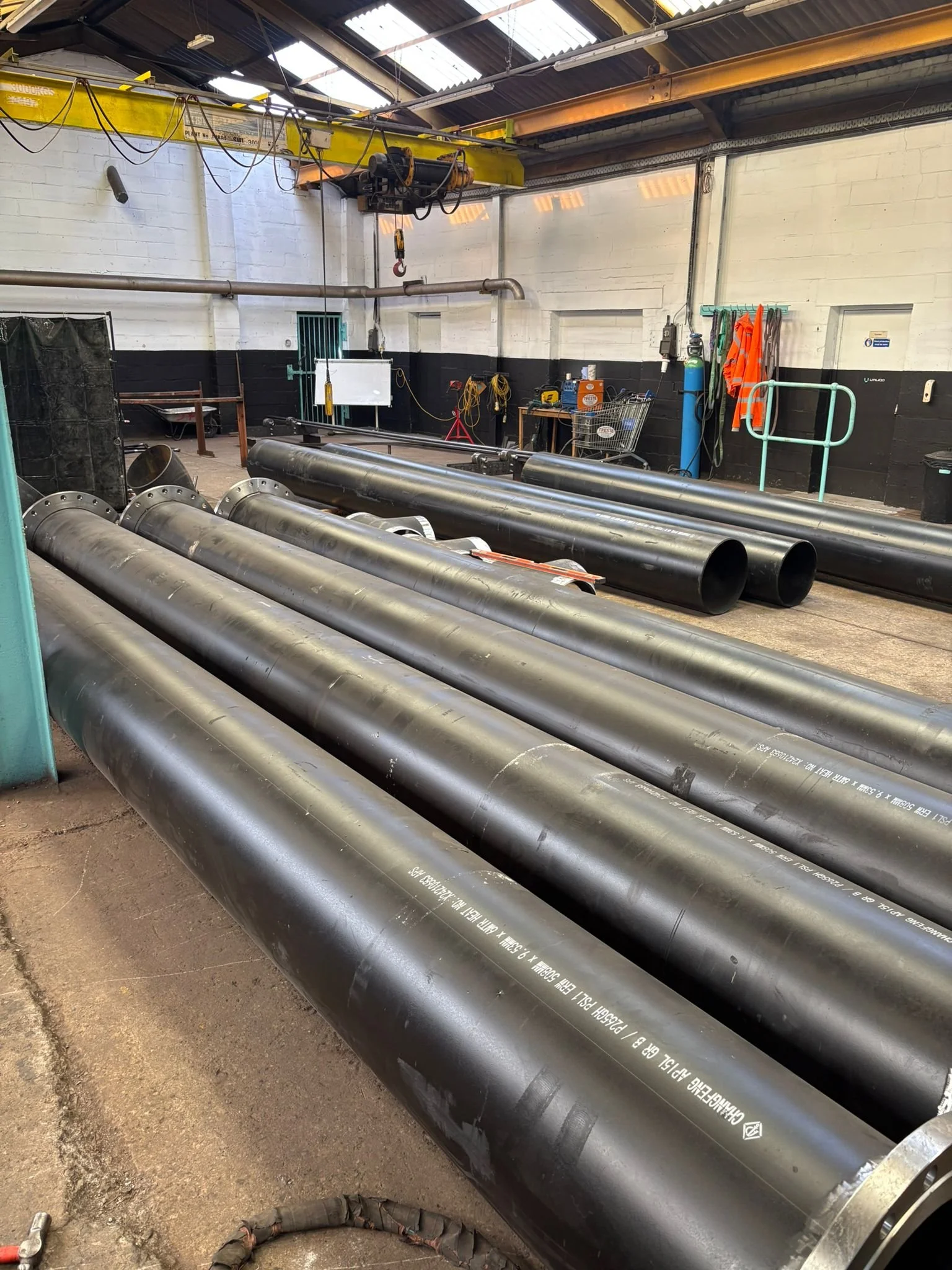 A workshop with large black pipes laid on the floor, some tools, safety equipment, and a yellow overhead crane system for lifting heavy objects.