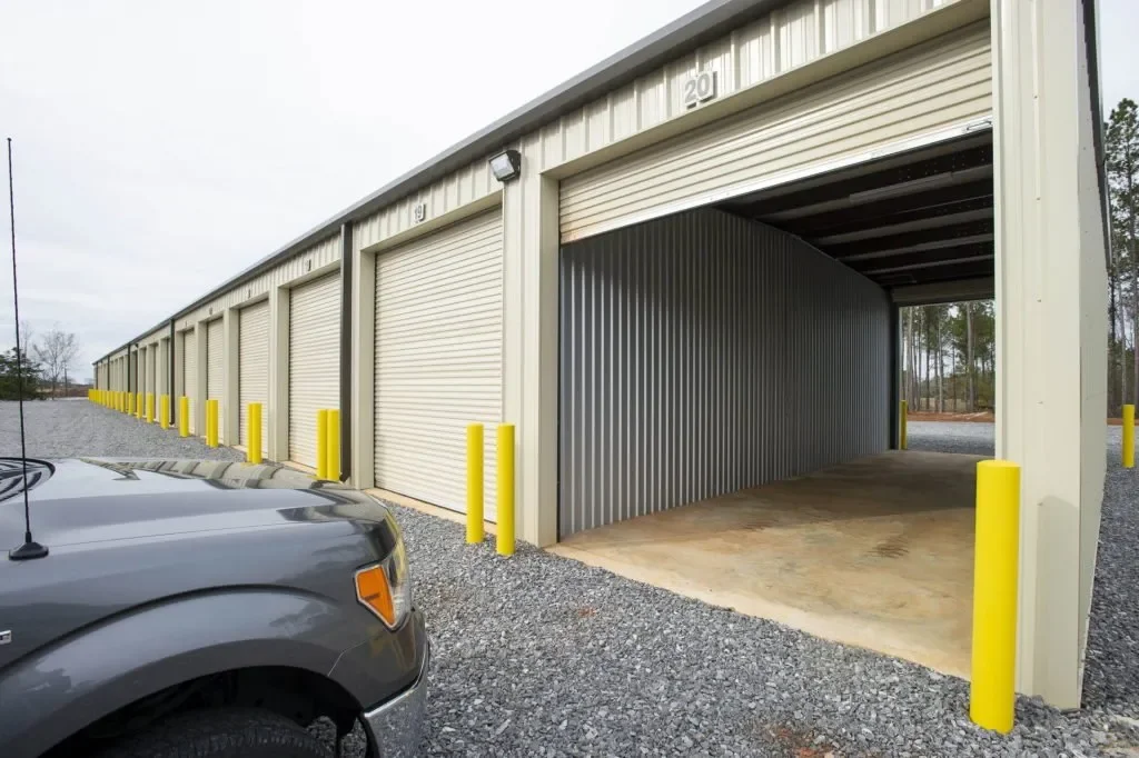 Row of storage units with closed and open doors, yellow safety bollards, and a gray vehicle in the parking lot.