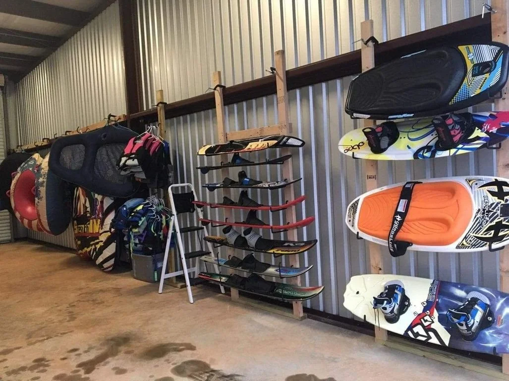 Display of wakeboards and water skis mounted on a wall with a metal siding background in a storage or shop area.
