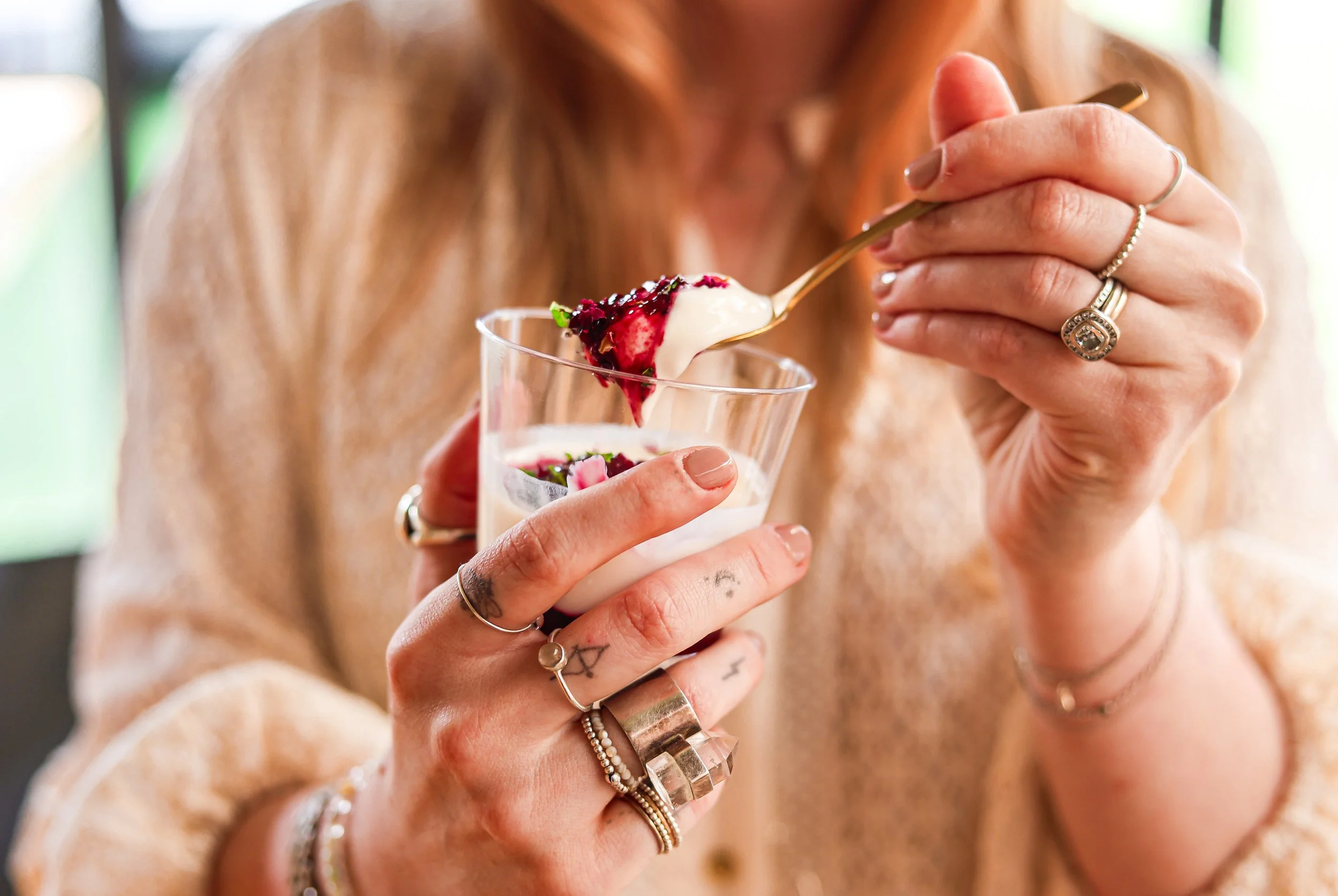 A woman with multiple rings and bracelets is holding a glass of dessert and spooning a spoonful of creamy dessert topped with berries.