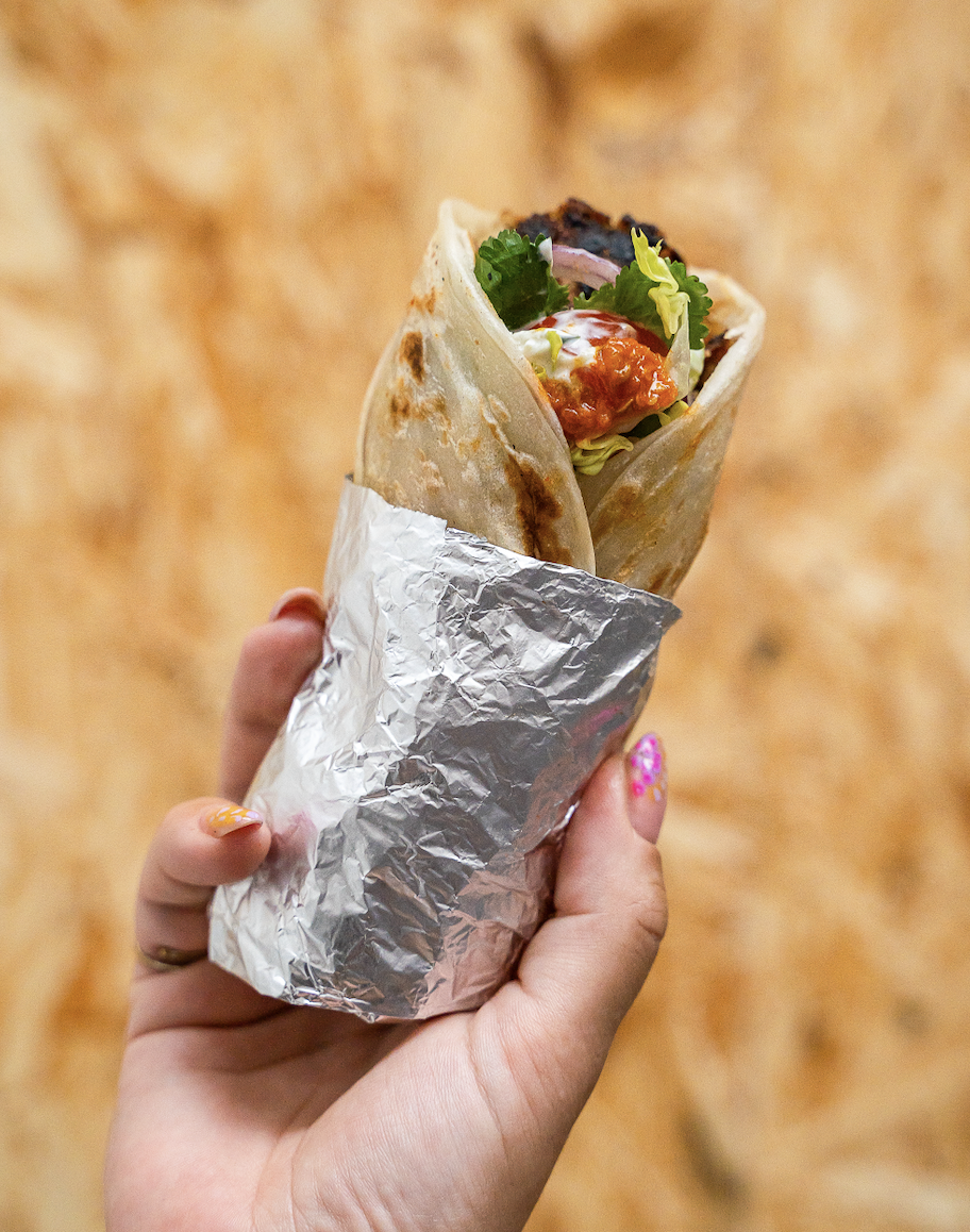 Close-up of a hand holding a partially wrapped burrito filled with lettuce, salsa, and other ingredients, against a blurred brown background.