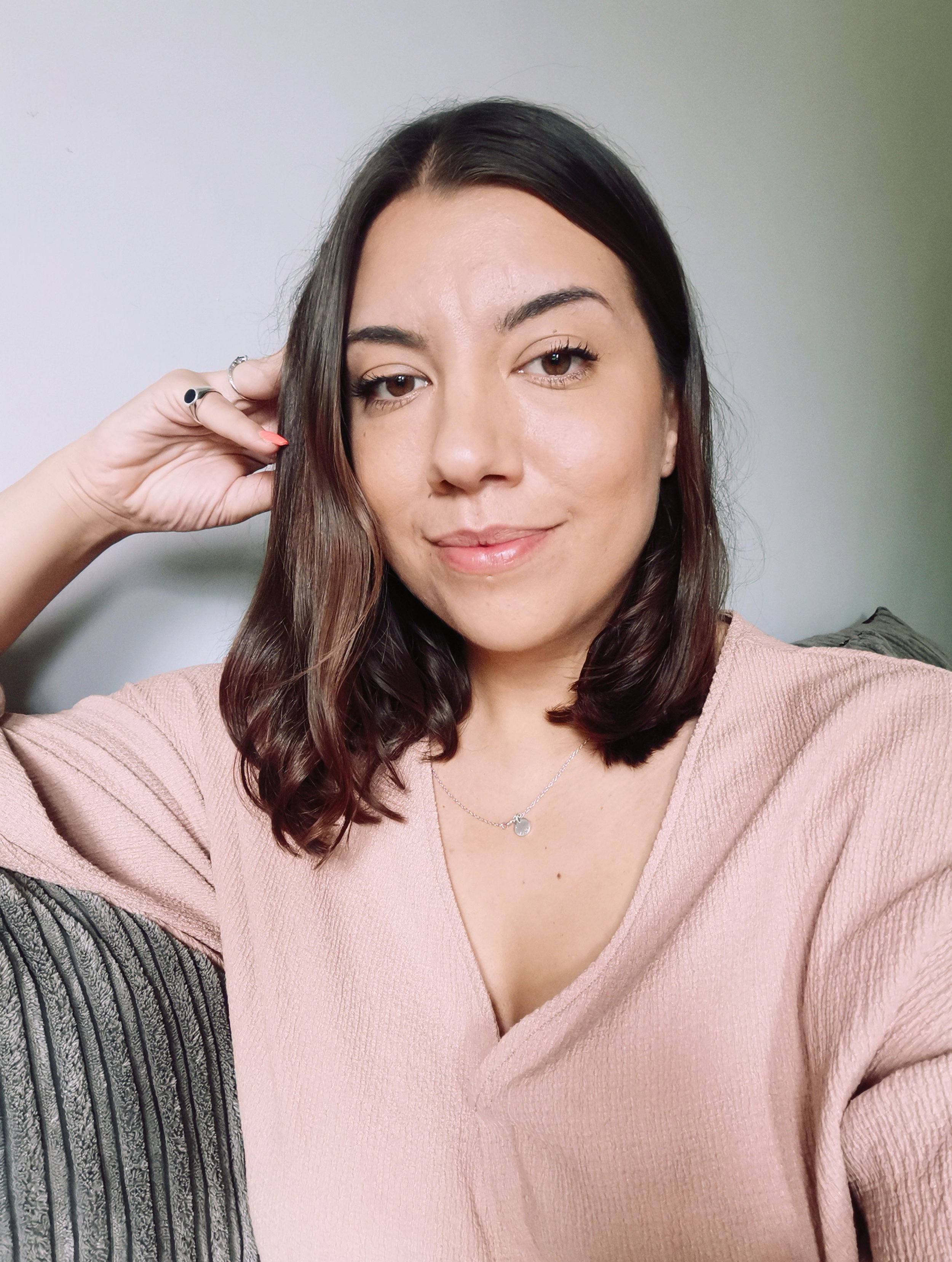 A woman with shoulder-length dark brown hair styled in loose curls, wearing a pink V-neck sweater, a delicate necklace with a small charm, and a black ring with a small colored design, smiling and touching her hair with her left hand against a plain light-colored wall.