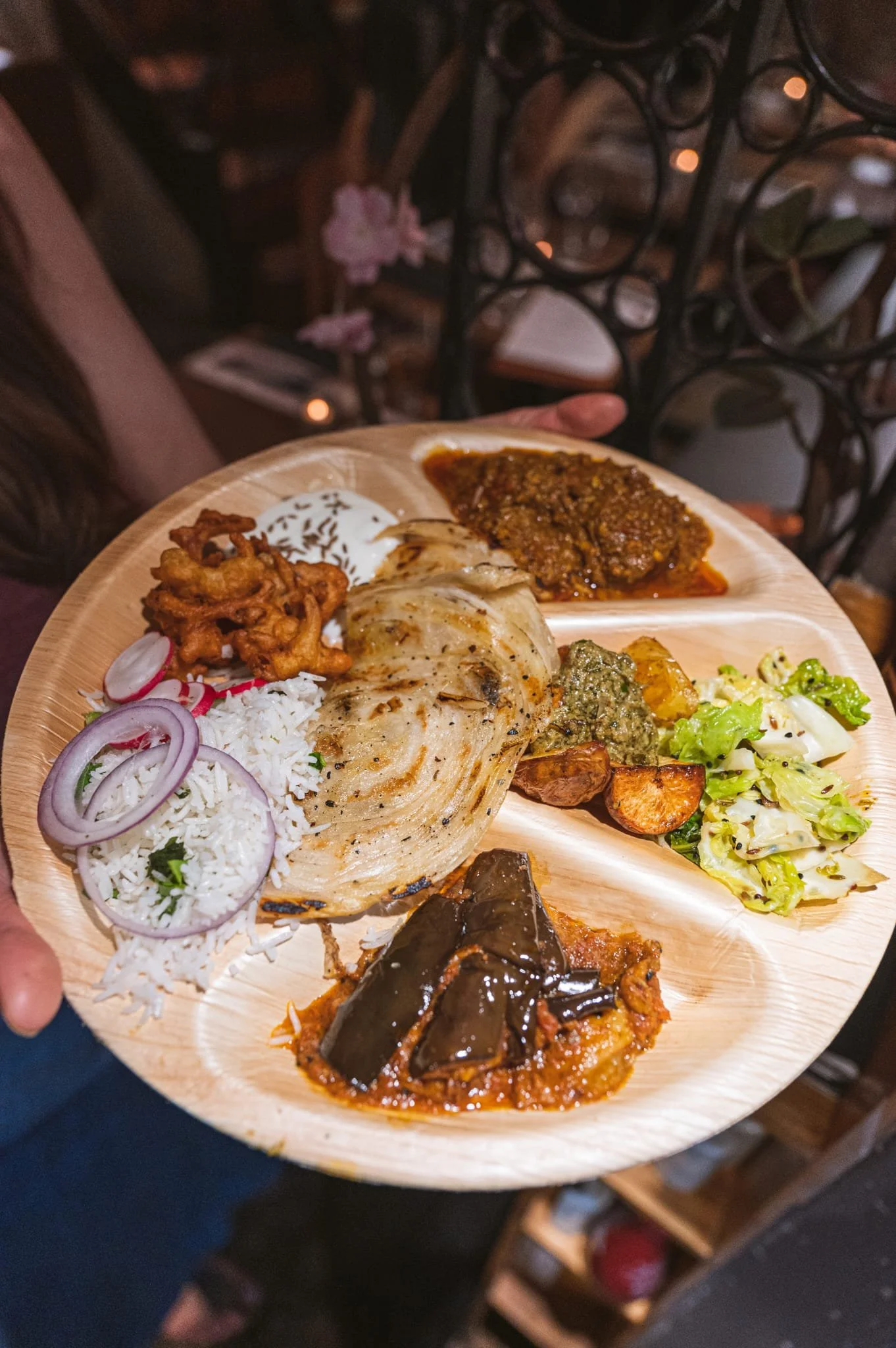 A wooden plate with various Indian dishes, including rice, grilled chicken, eggplant curry, vegetable salad, and meat curry, held by a person with a dark background.