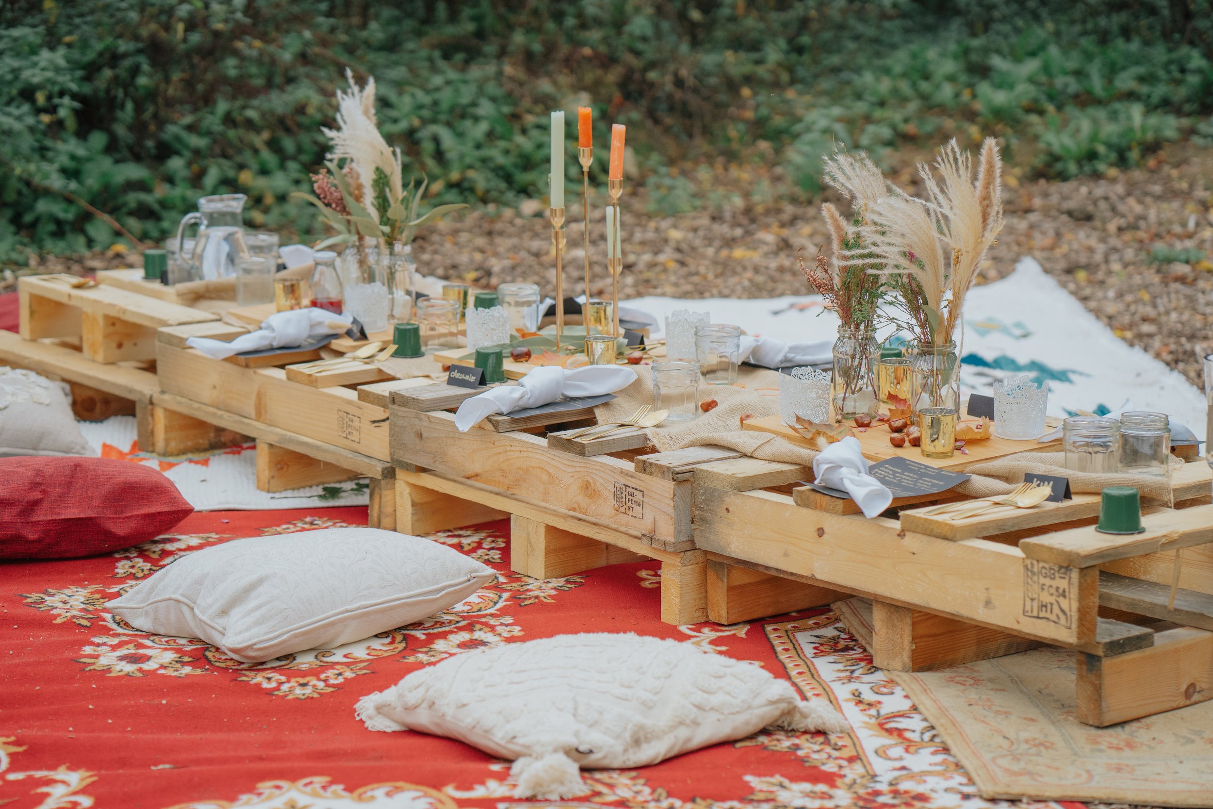 A rustic outdoor dining setup featuring a wooden pallet table on a decorative red and cream rug with white and red pillows for seats. The table is set with glassware, white napkins, candles, and floral centerpieces with dried pampas grass and greenery. The scene is outdoors with green foliage in the background.