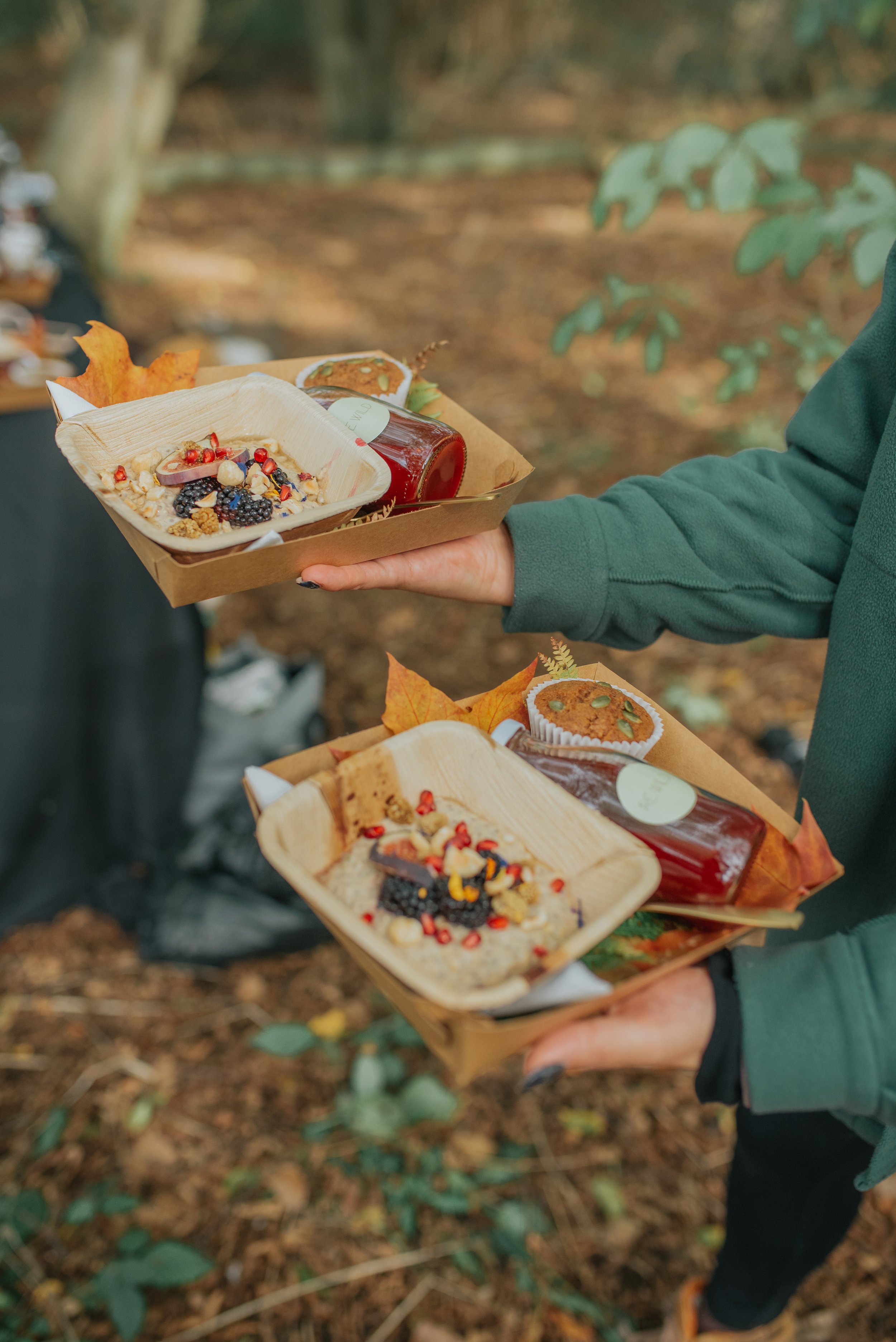 Two people holding trays of snacks outdoors, with a background of trees and fallen leaves.