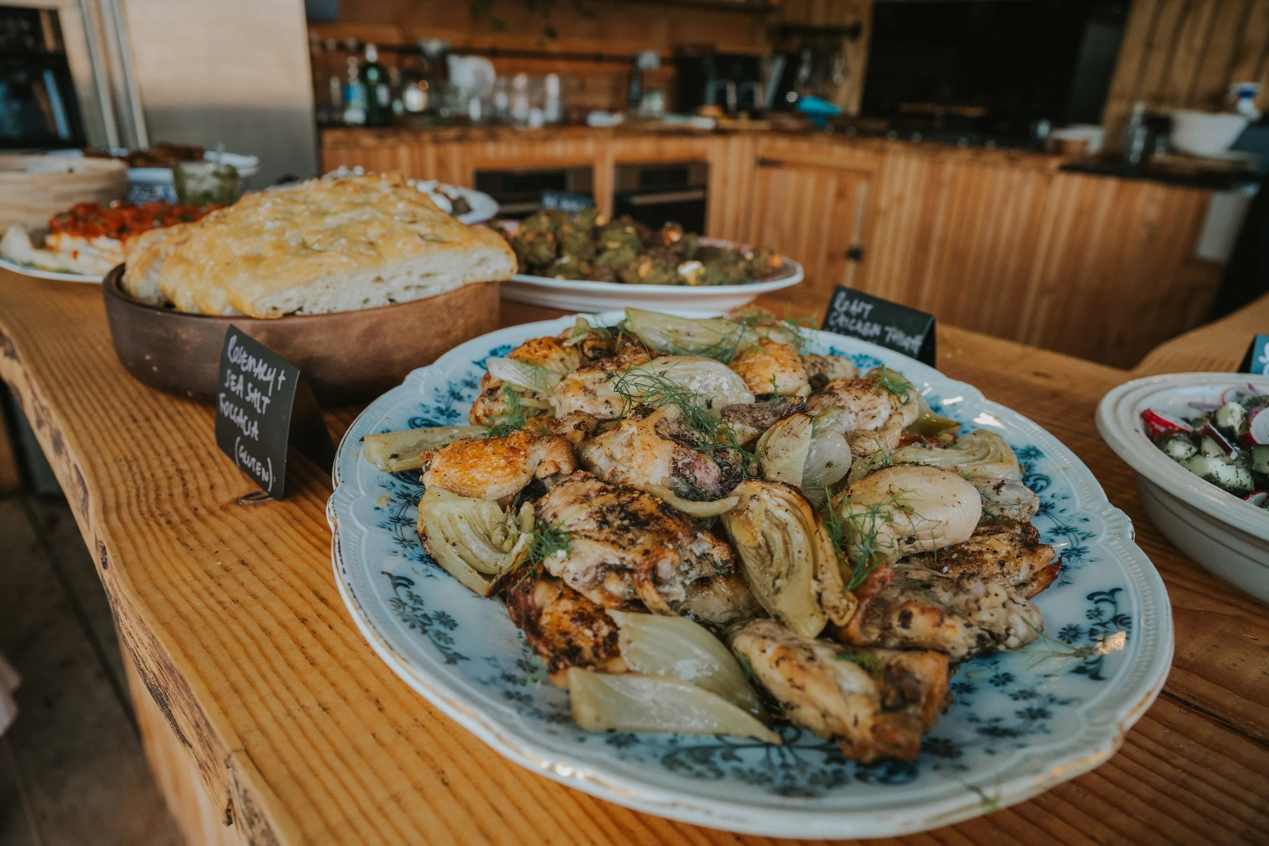 A wooden table displaying various plates of food, including a plate of grilled chicken with onions and herbs, a large bowl of bread or focaccia, and a plate of salad with radishes and cucumbers, in a cozy rustic kitchen setting.