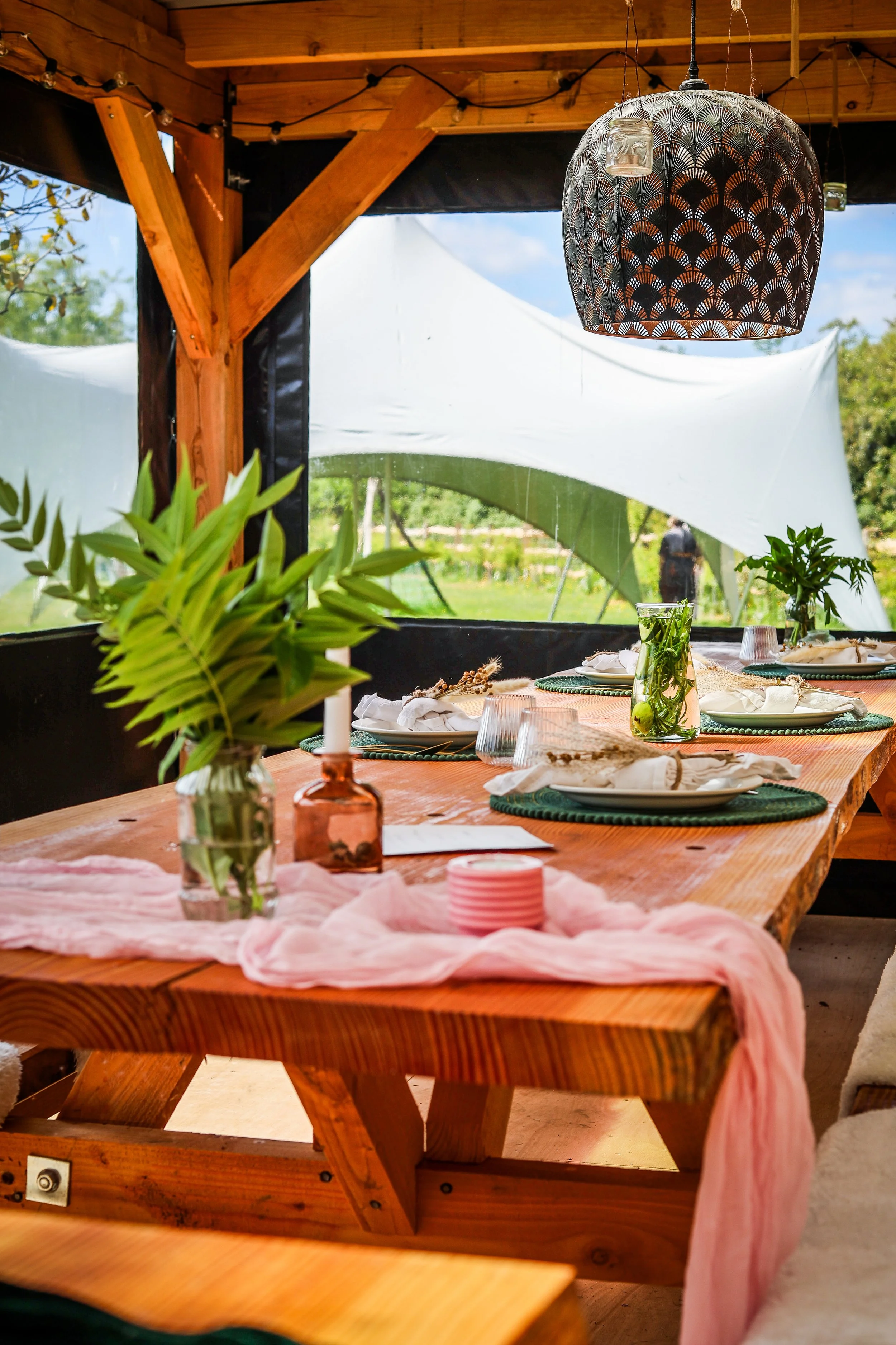 A decorated outdoor dining table with plates, glasses, and tableware, set under a wooden structure with a decorative hanging lamp and green plants, overlooking a garden.
