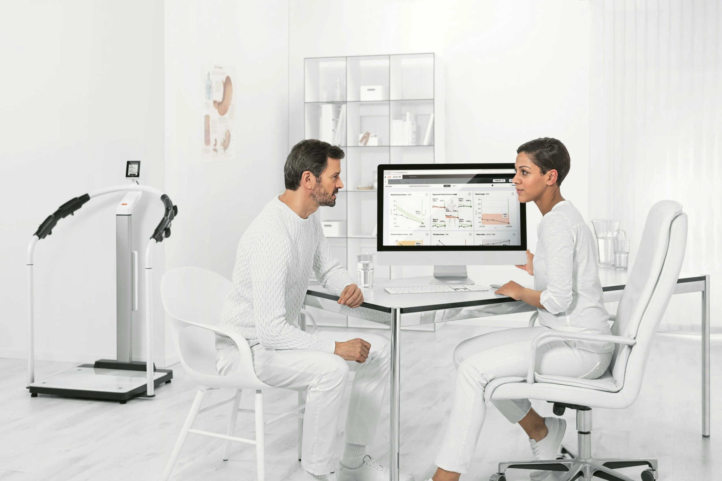 A patient consulting a healthcare professional in a medical office, with medical equipment and a computer displaying graphs on the desk.