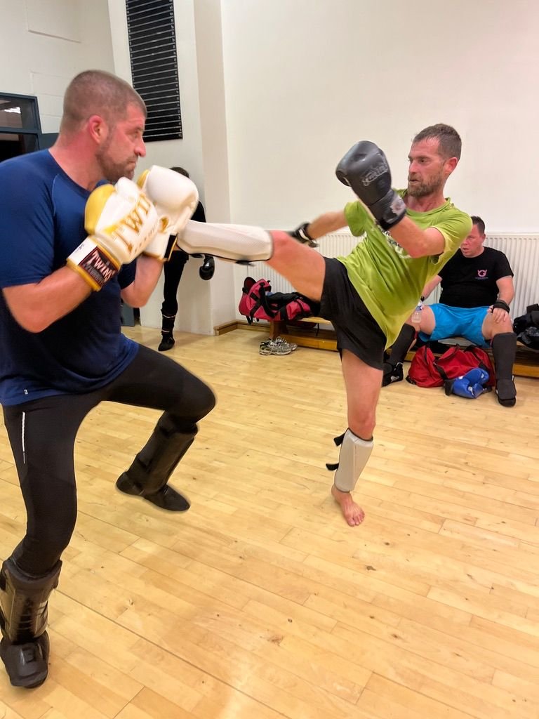 Deux hommes pratiquent la boxe en salle d'entraînement, avec un homme en vert effectuant un coup de pied sous la garde d'un autre homme en bleu.